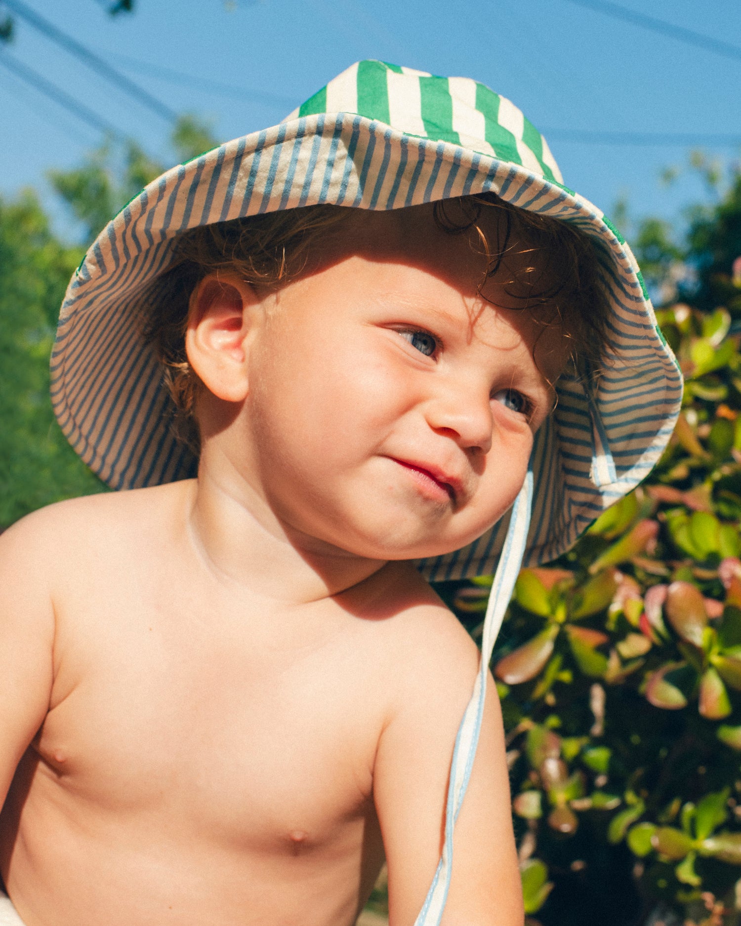 Close up of toddler model is wearing Big Reversible Sun Hat in Blue Pinstripe in a sunny lush area.