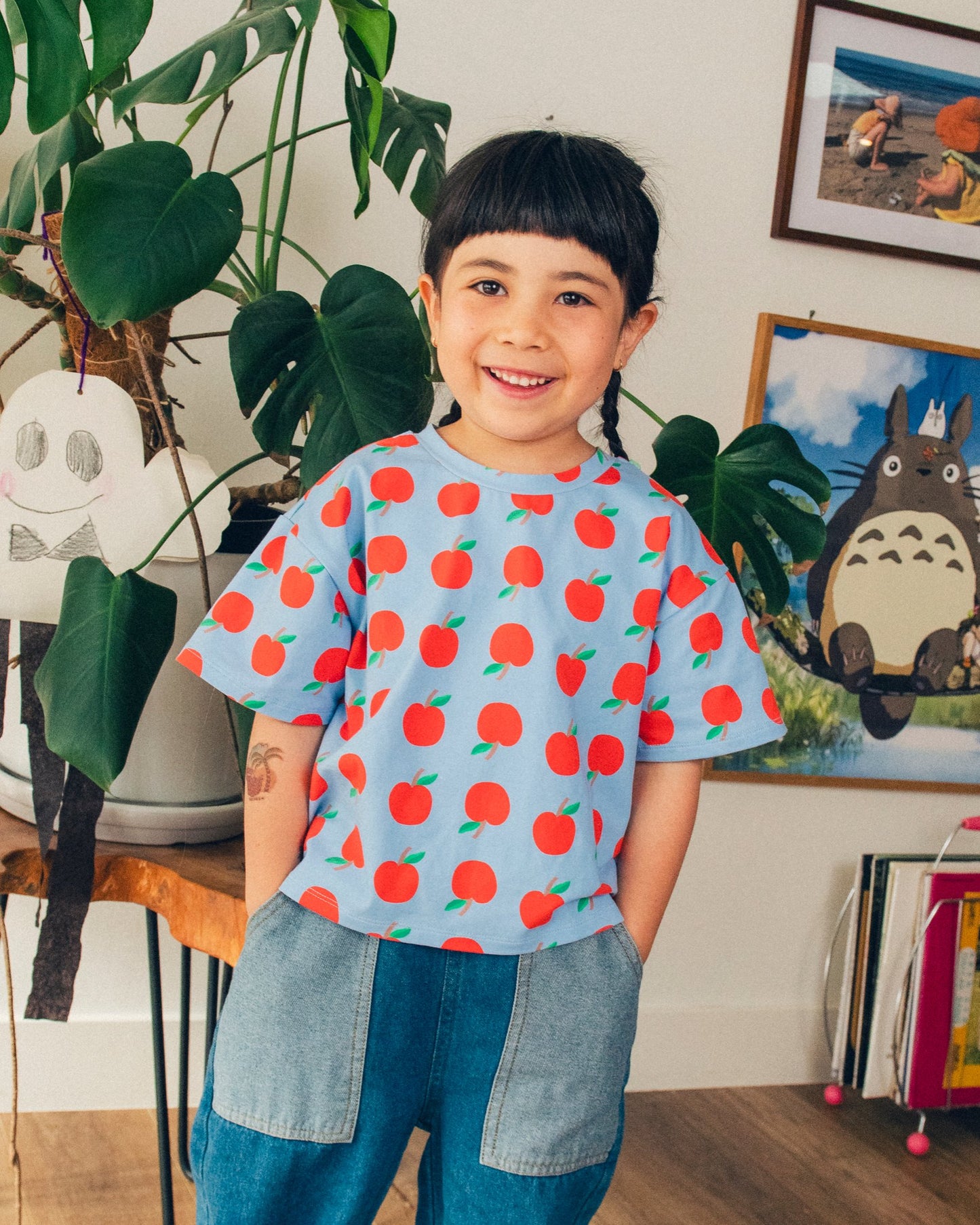 Model is wearing Graphic Tee in Blue Apple and jeans in a living room setting.@model_info:Zoe is 6 years old and wearing 4T.
