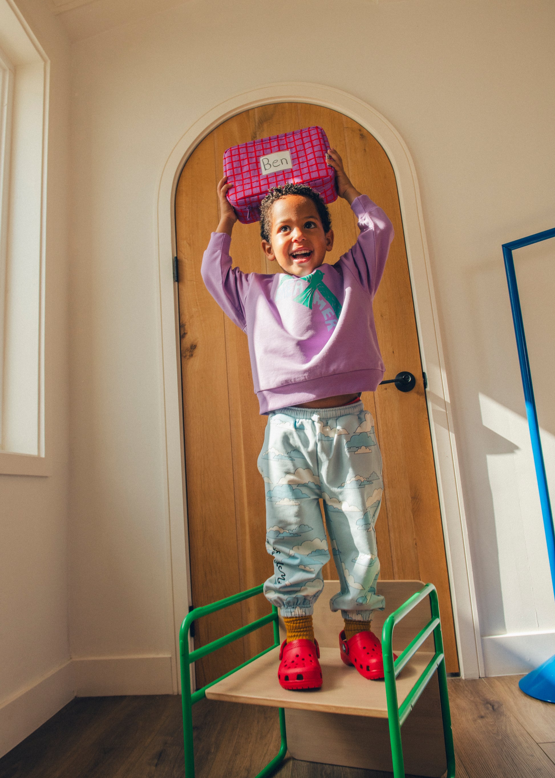 Model is holding Cool Kid Lunch Box in Orchid Grid on top of head and wearing French Terry Jogger in Blue Clouds and Graphic Sweatshirt in Purple Bird in front of door in sunlit room. @model_info:Zion is 3 years old and wearing 4T.