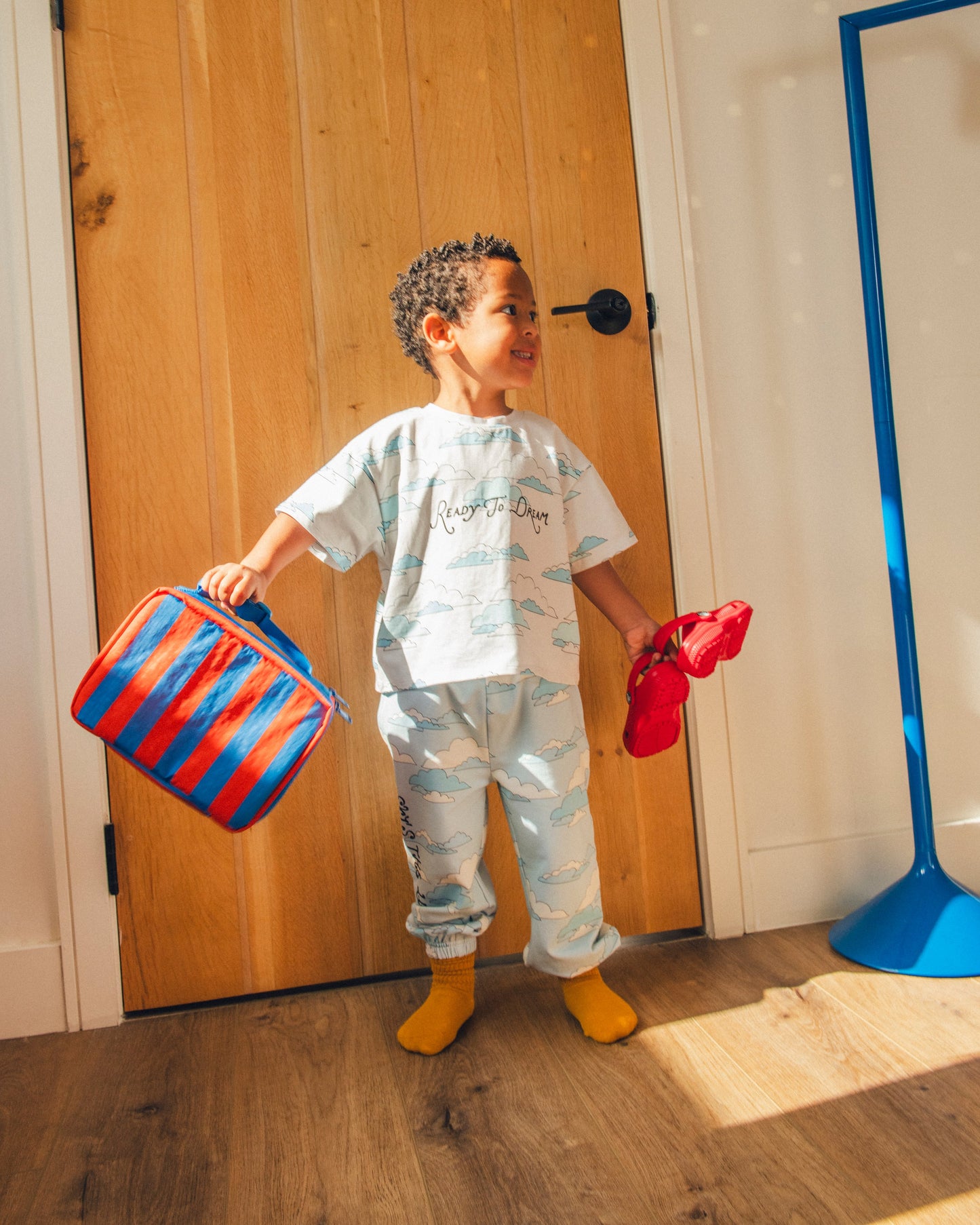 Model is standing at front door holding  shoes and Cool Kid Lunch Box in Blue & Rust Stripe and wearing French Terry Jogger in Blue Clouds and Graphic Tee in Ivory Clouds, ready for the day.@model_info:Zion is 3 years old and wearing 4T.