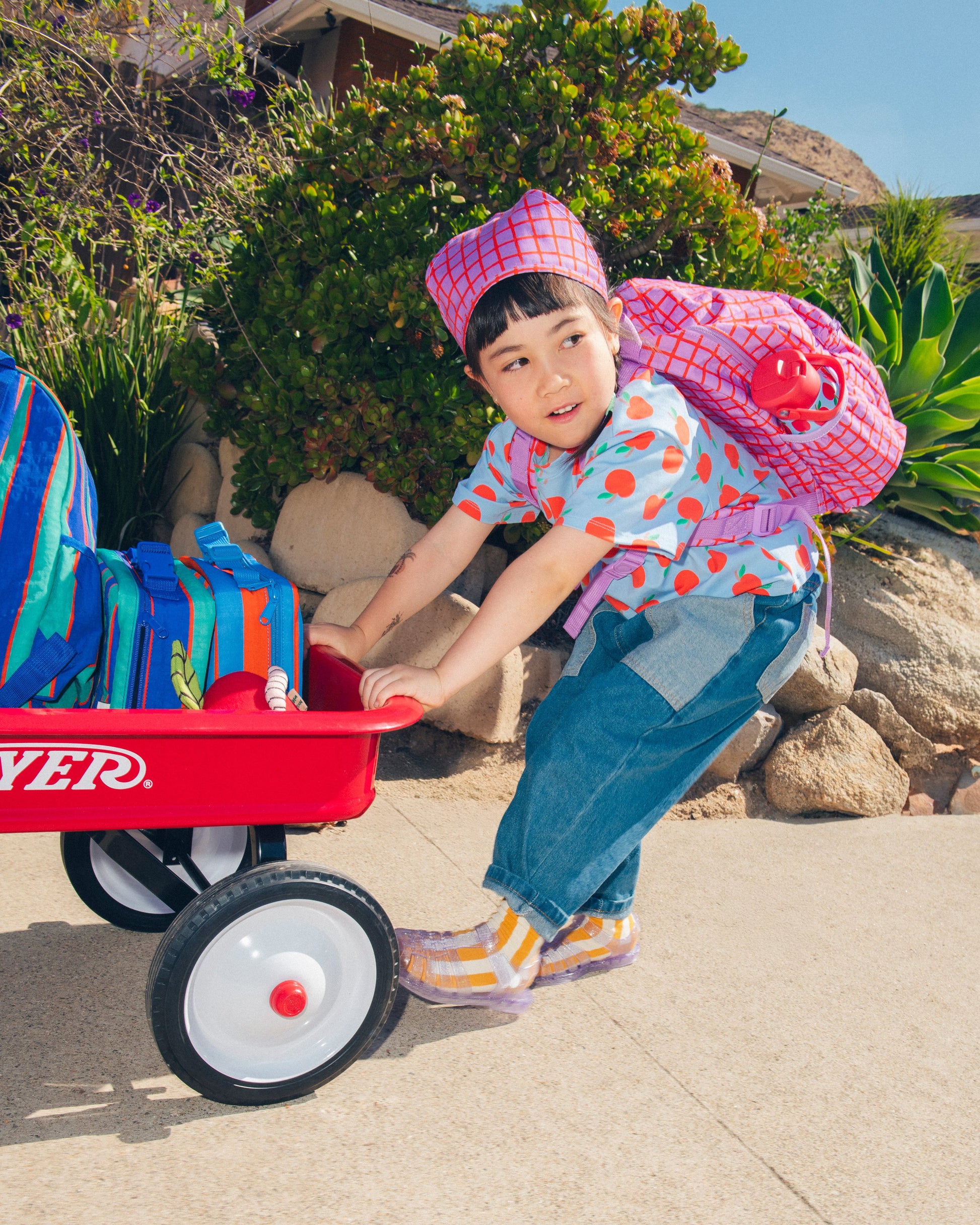 Child pushing a red wagon with Cool Kid Backpack in Navy & Green Stripe, Cool Kid  Lunch Box in Navy & Green Stripe and Blue & Rust Stripe, while wearing Cool Kid Backpack in Orchid Grid, alongside a sidewalk in a neighborhood.