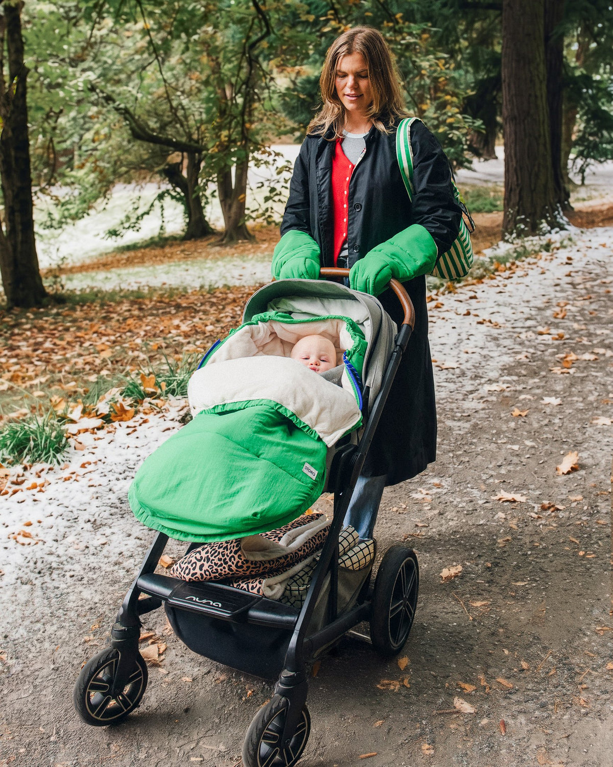 Mother is pushing baby in grey stroller through a park with snow on the ground. Baby is snuggled into the Cozy Stroller Footmuff in Green and the mother is wearing Cozy Stroller Mitts in Green, jeans, full black coat and Stroller Party™ Bucket Bag in Green Stripe.
