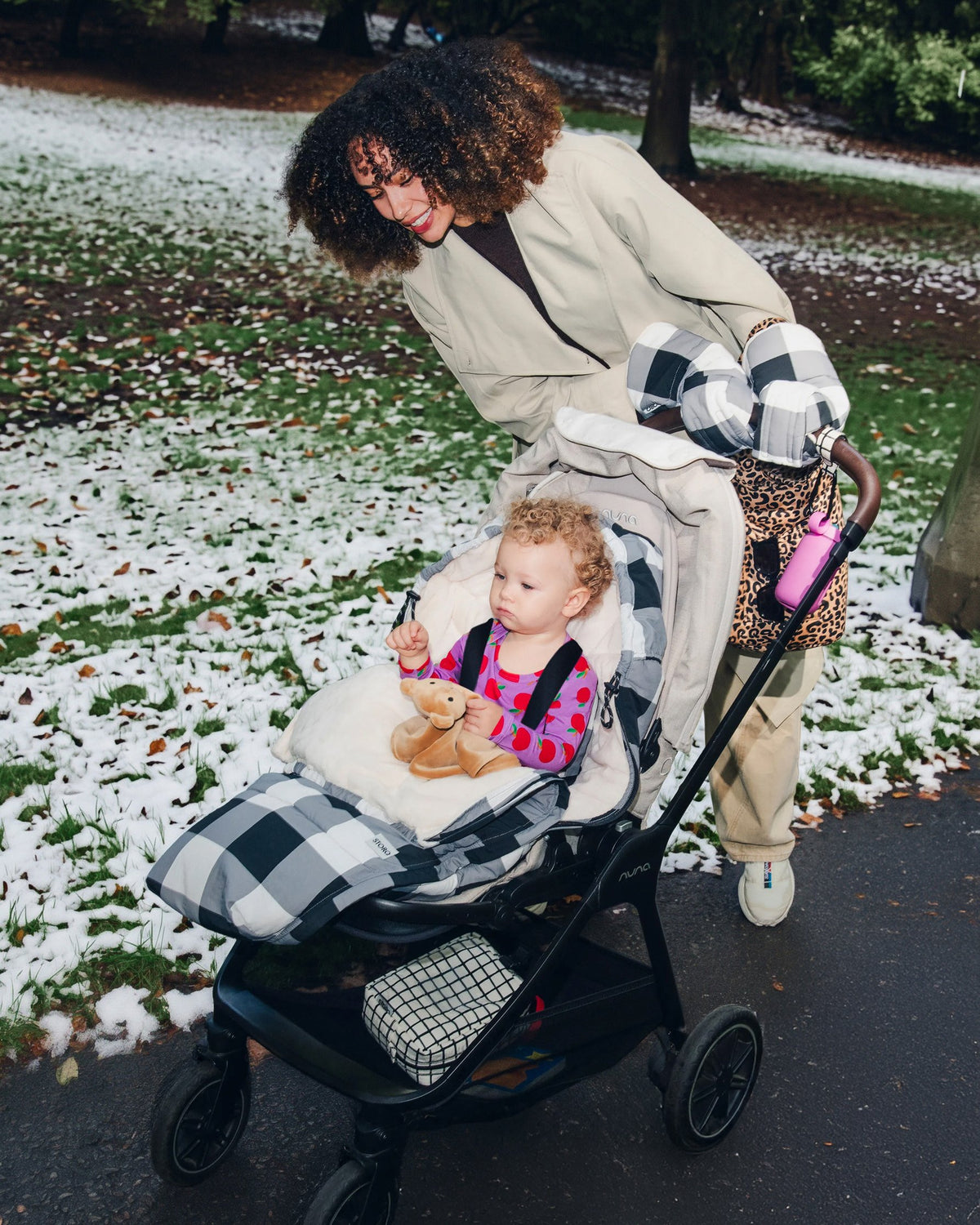 A woman pushes a stroller with a young child in a snowy winter outdoor park setting. The stroller is covered in Cozy Stroller Footmuff in Buffalo Plaid and the woman is wearing matching Cozy Stroller Mitts in Buffalo Plaid, and the baby is wearing Long Sleeve Bamboo Onesie Two-Pack in Apple.