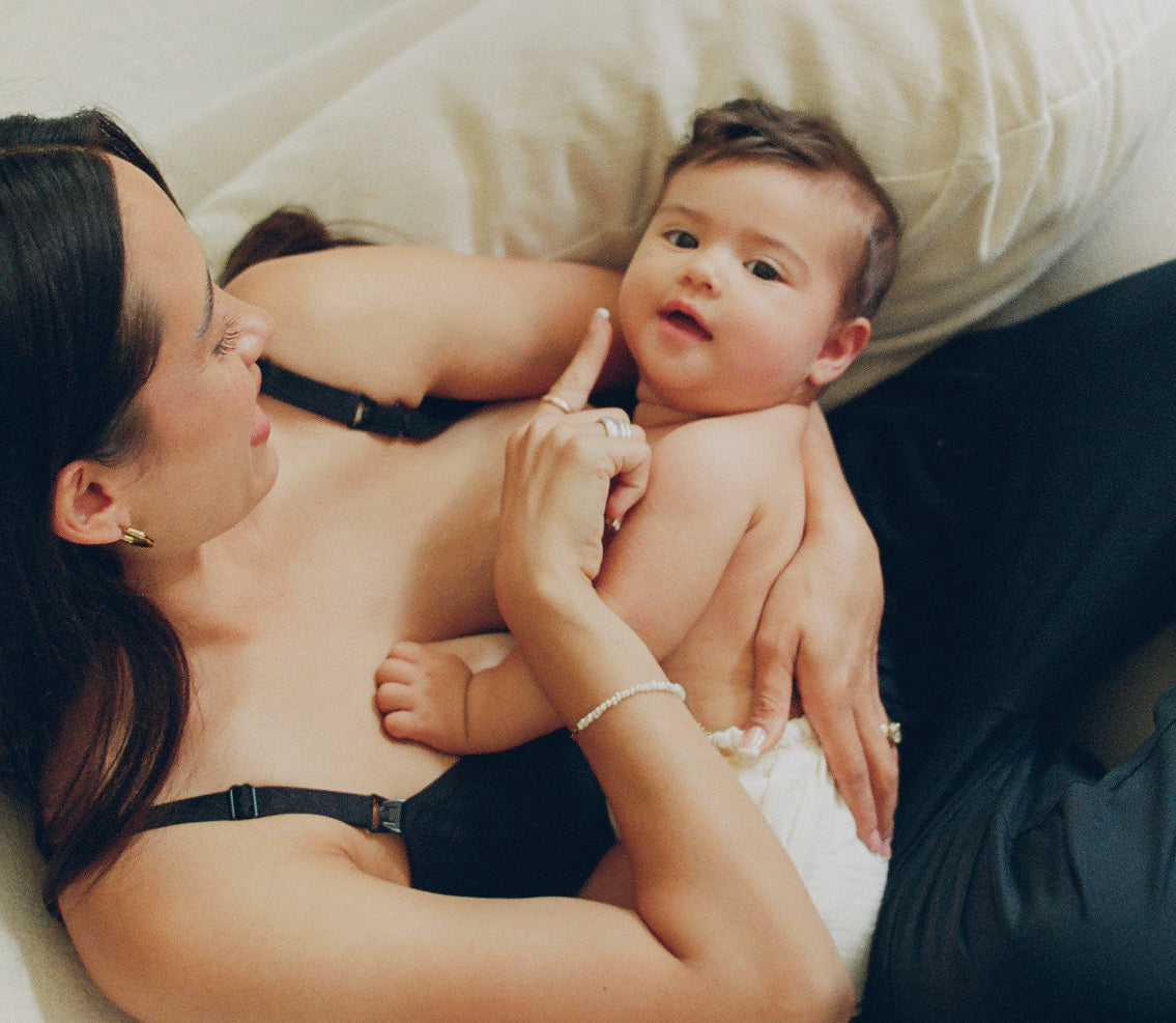 Woman is reclining on white couch while breastfeeding baby, who is looking up at the camera, in a light filled room.
