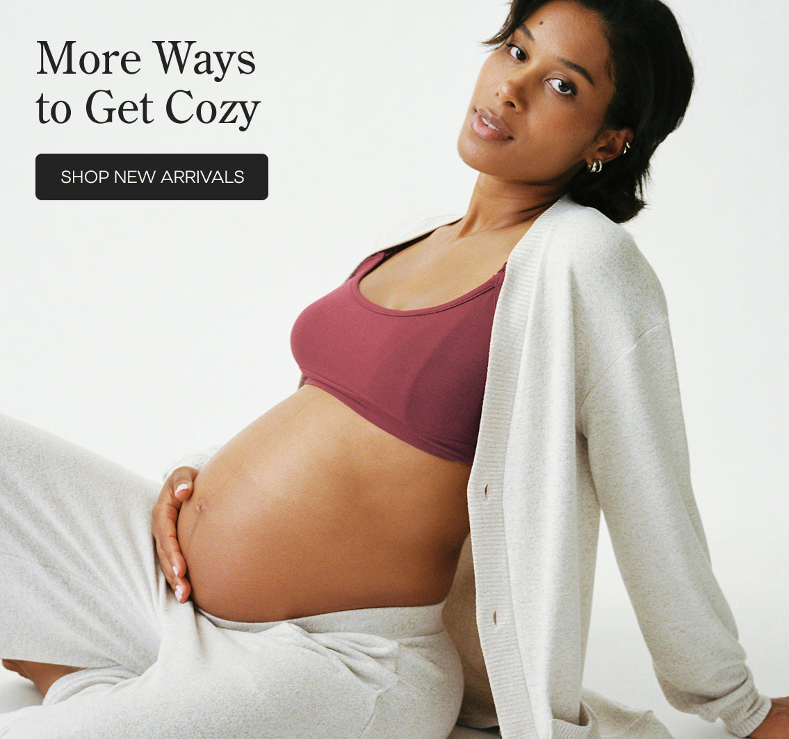Model is reclining and wearing CozyKnit Wide Leg Lounge Pant in Oat, and CozyKnit Long Cardigan in Oat, and French Terry Maternity & Nursing Bralette in Deep Burgundy on a beige background. Black text on the left reads 'More Ways to Get Cozy' with a 'SHOP NEW ARRIVALS' button.