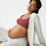 Model is reclining and wearing CozyKnit Wide Leg Lounge Pant in Oat, and CozyKnit Long Cardigan in Oat, and French Terry Maternity & Nursing Bralette in Deep Burgundy on a beige background.