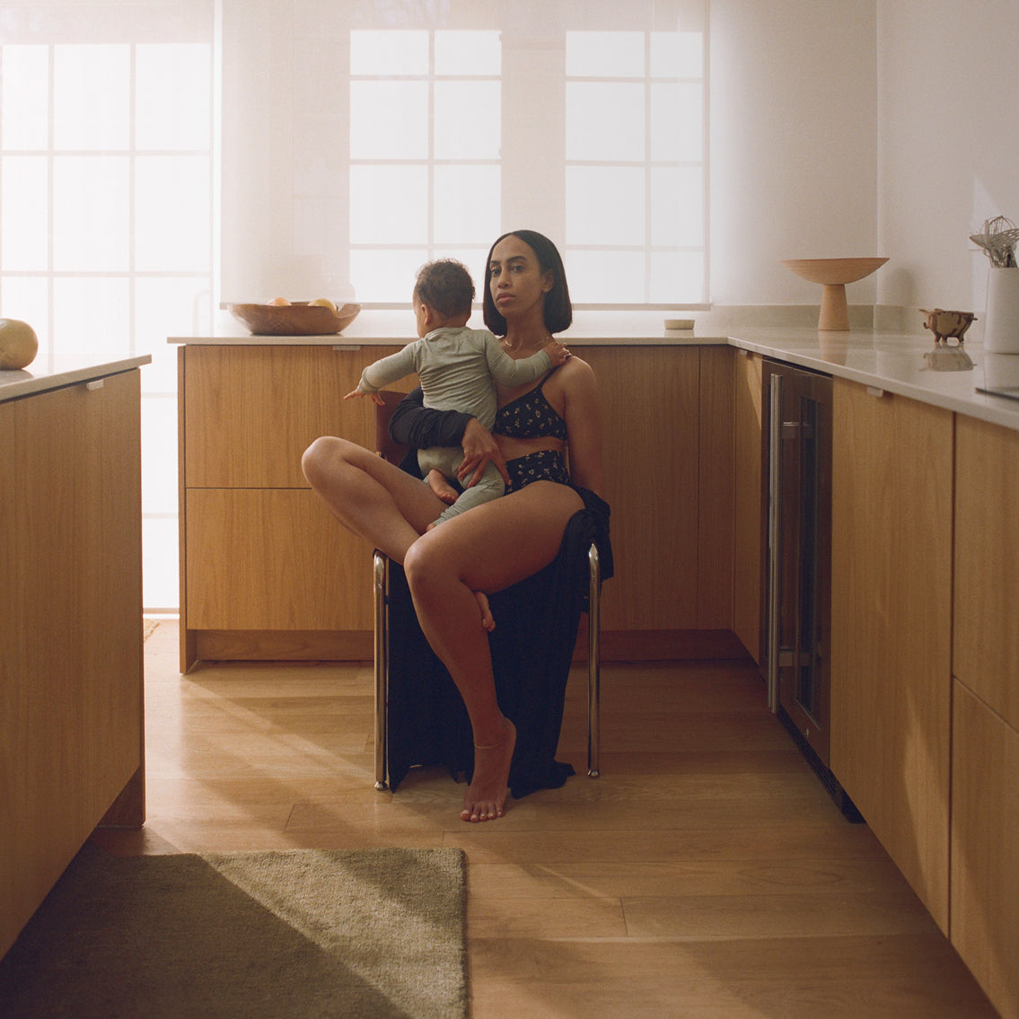 Baby sitting in mother's lap on chair in modern kitchen wearing Every Stage Triangle Bralette and Brief in Black Wildflower, with light filled windows in background.
