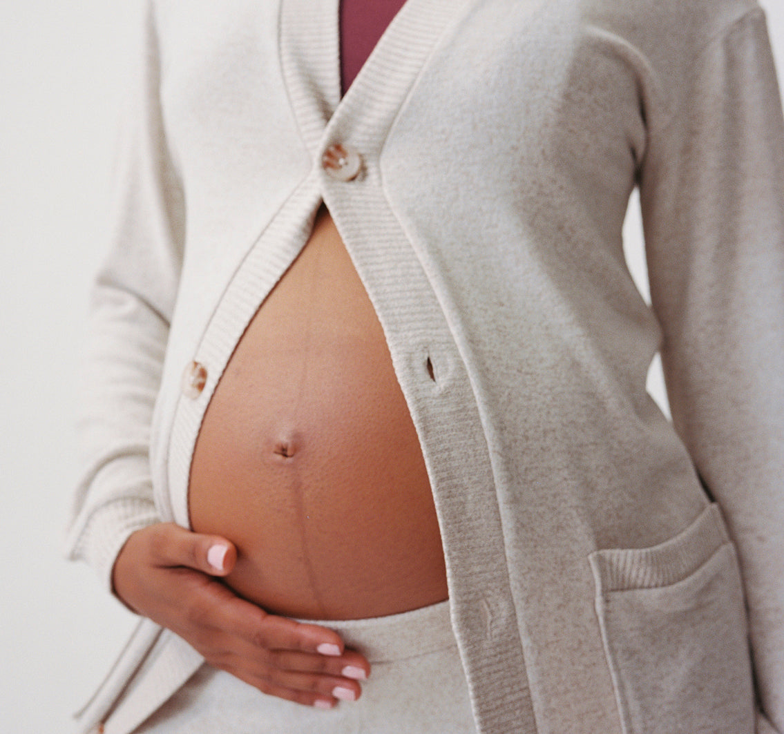 Close up of model wearing CozyKnit Wide Leg Lounge Pant in Oat, and CozyKnit Long Cardigan in Oat, and French Terry Maternity & Nursing Bralette in Deep Burgundy on a beige background.