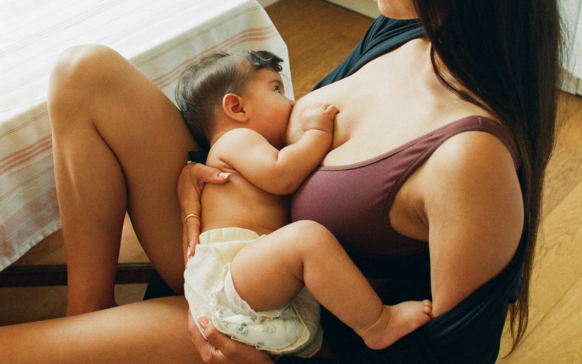 Mother breastfeeding baby while wearing Every Stage Crossover Bra in Cocoa and reclining in chair at table in a dining room setting.