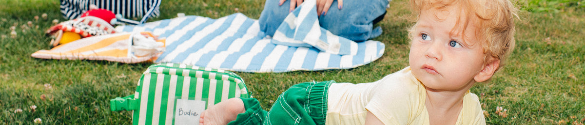 Toddler sitting outside by Inside/Outside Play Mat in Blue Wide Stripe, Packable Park Carryall in Navy Stripe and Jumbo Cooler Bag in Green Stripe, on the grass.