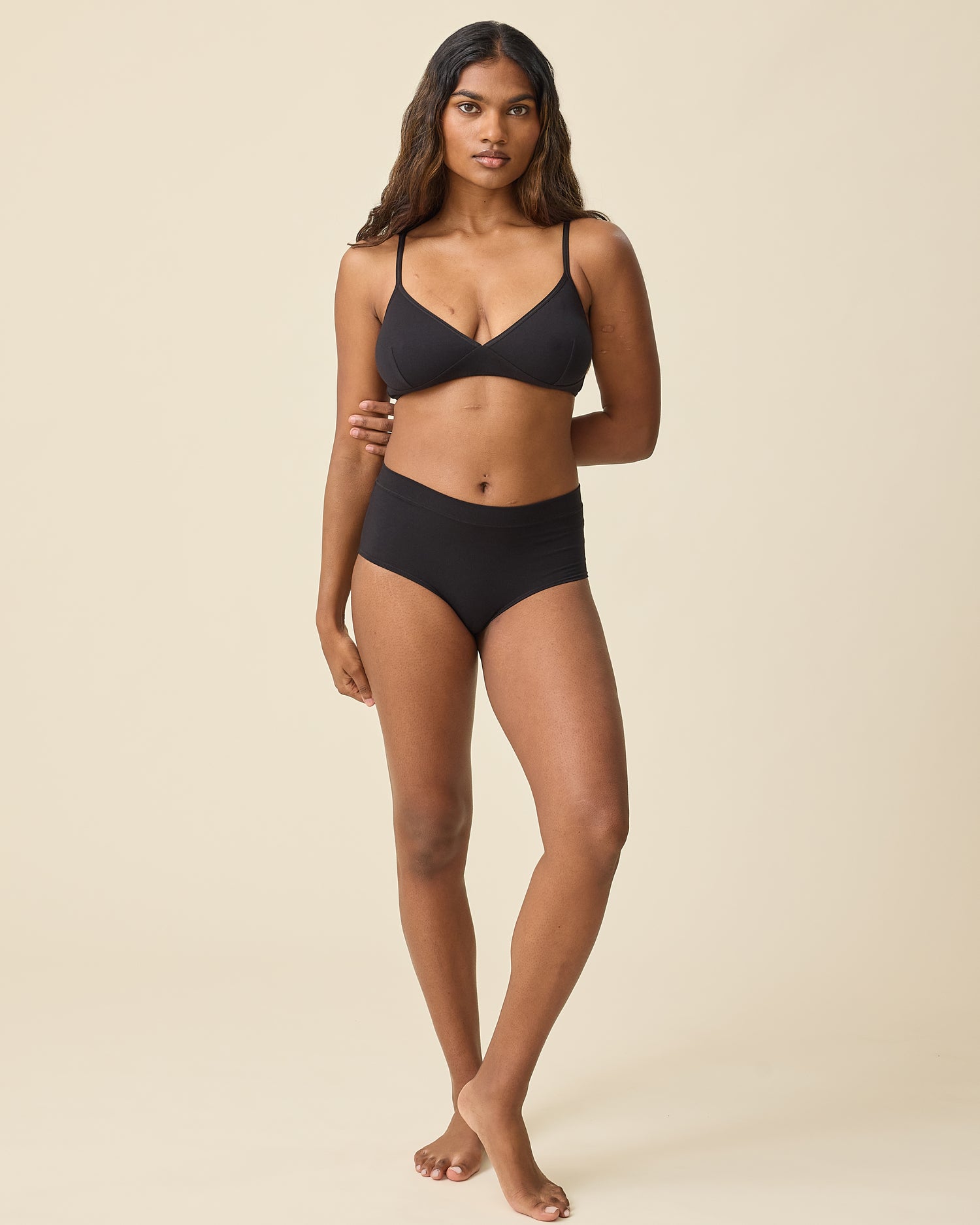 Model is wearing Every Stage Triangle Bralette in Black and Every Stage Brief in Black on a beige background.