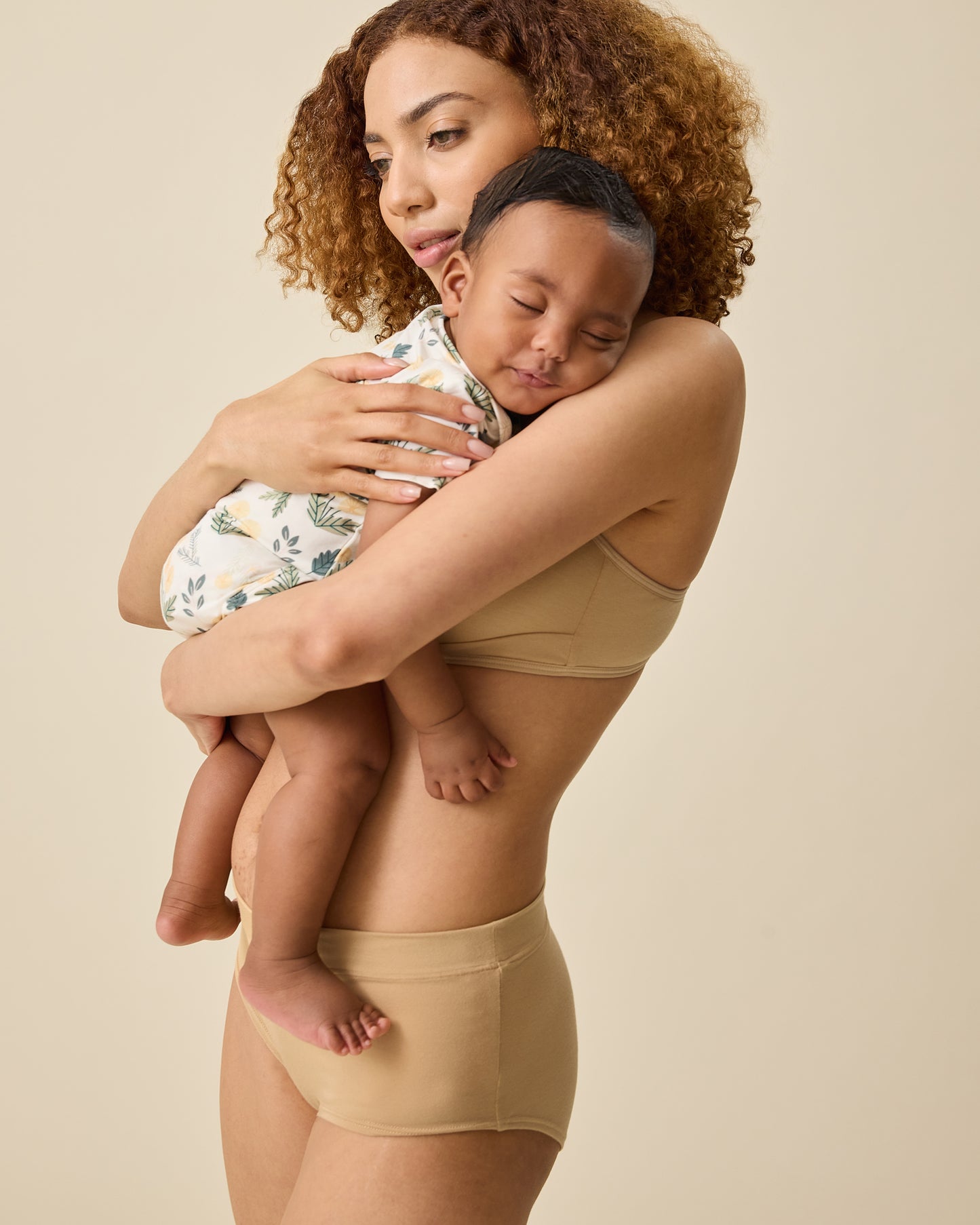 Side view of model holding a sleeping baby and wearing Every Stage Brief in Toffee and matching bra on a beige background