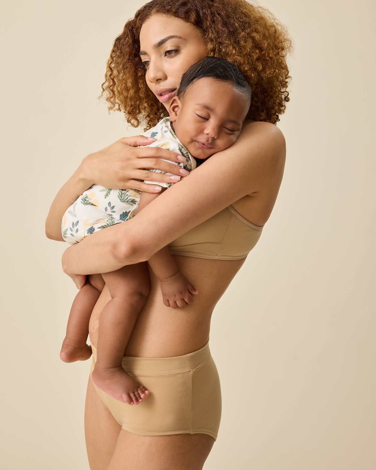 Side view of model holding a sleeping baby and wearing Every Stage Brief in Toffee and matching bra on a beige background