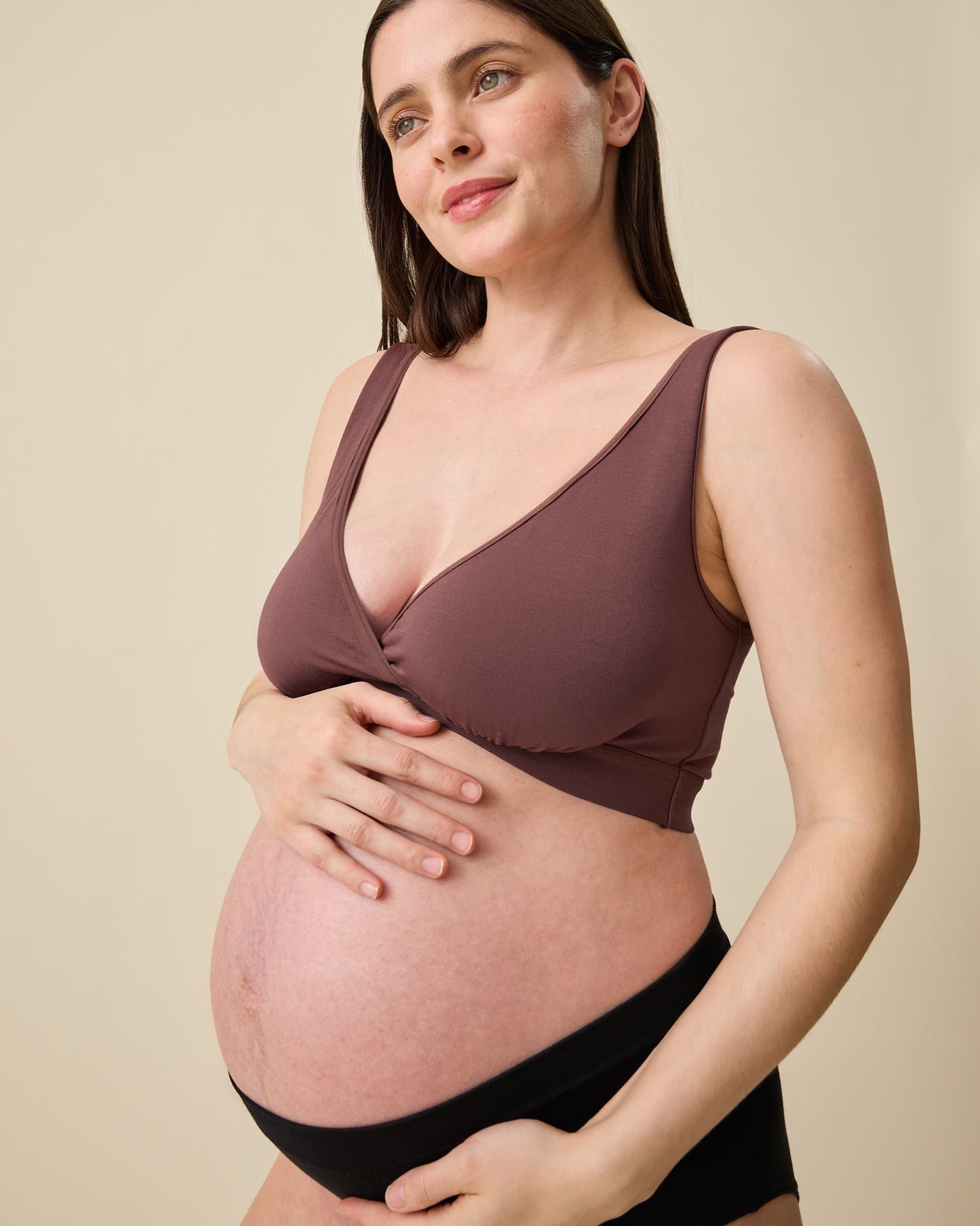 Pregnant model wearing Every Stage Crossover Bra in Cocoa and Every Stage Brief in Black on a beige background