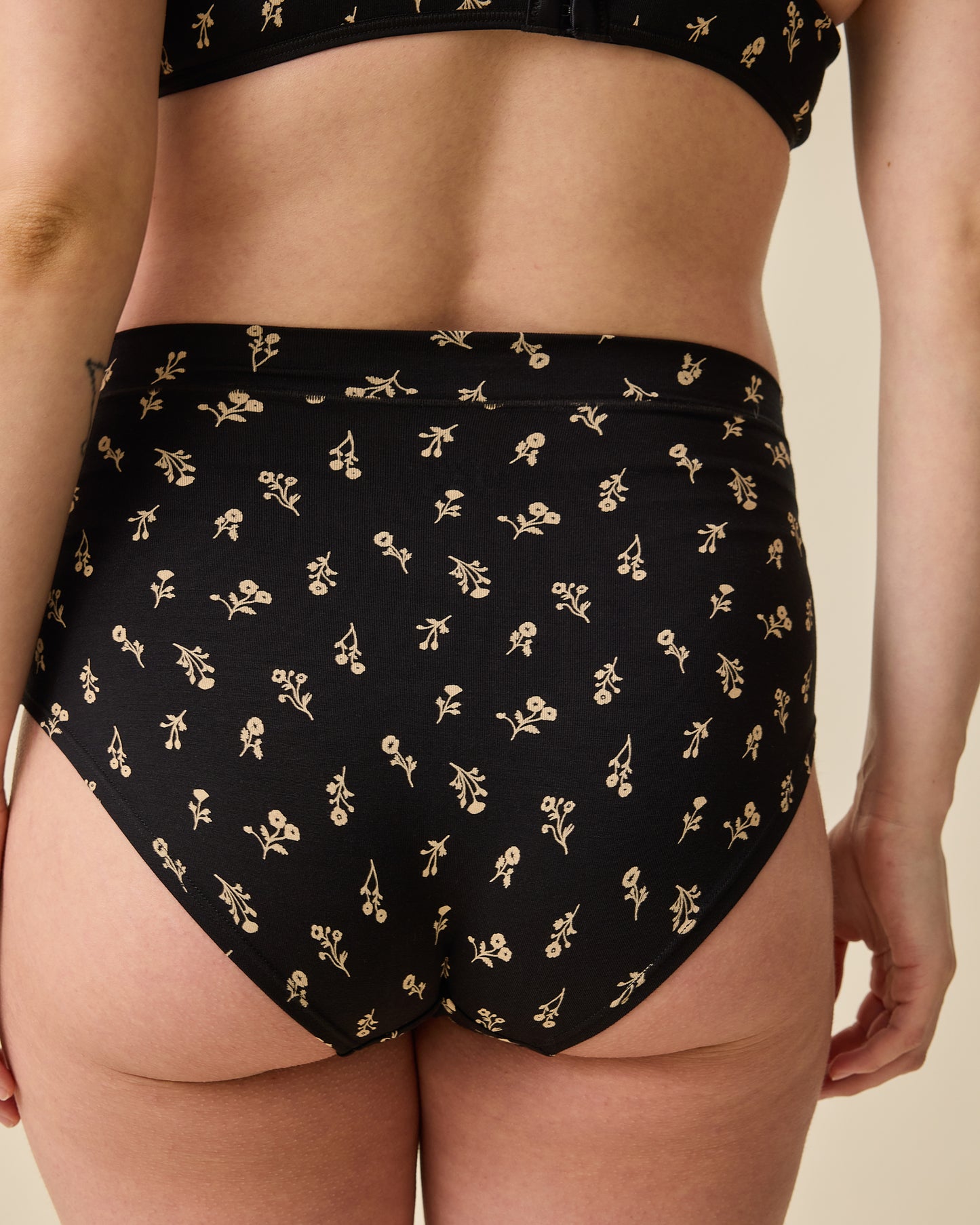 Back view of Model wearing Every Stage Triangle Bralette in Black Wildflower and Every Stage Brief in Black Wildflower on a beige background