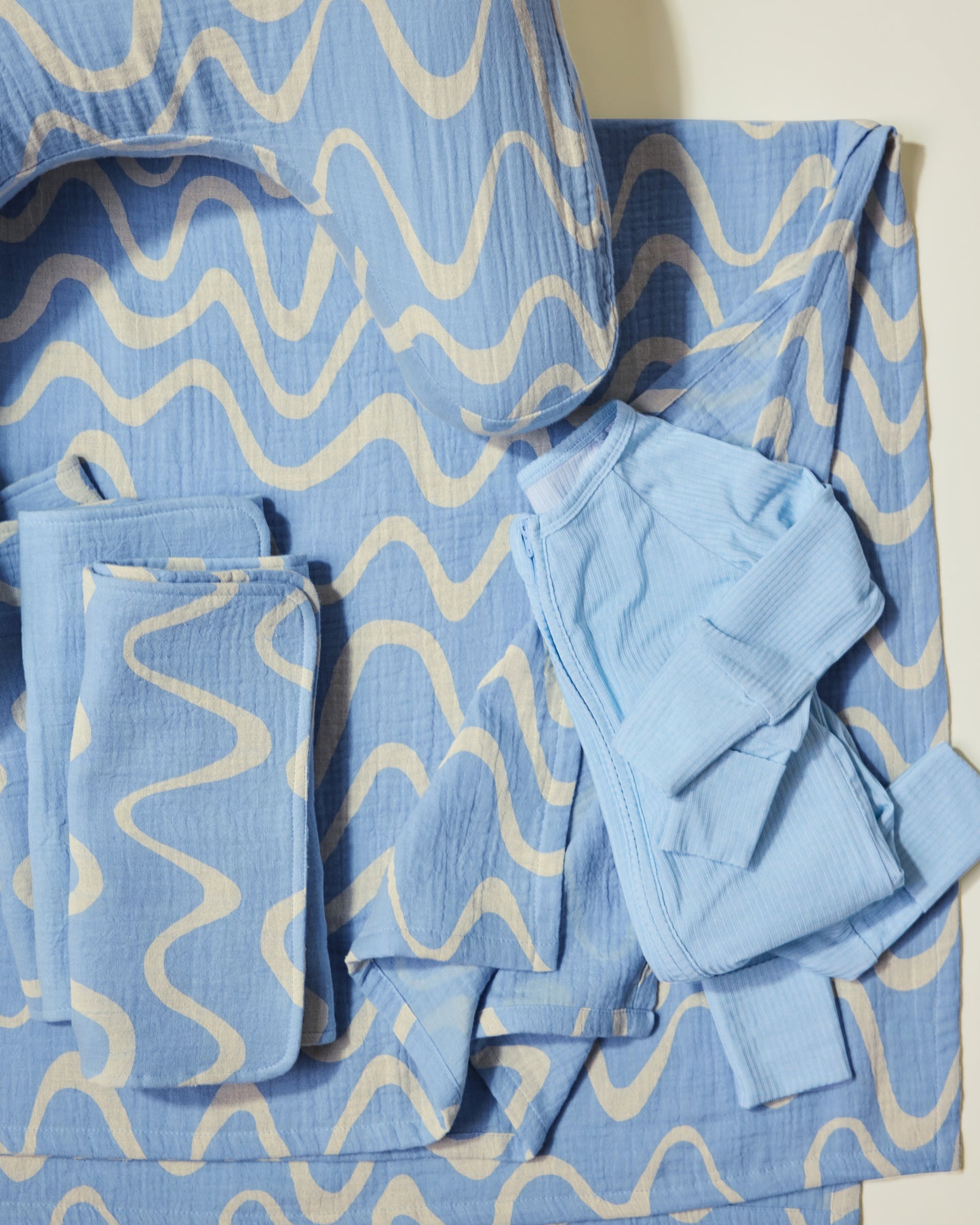Set of blue and white wave patterned nursing pillow, burp cloths, swaddle and baby onesie on a light background