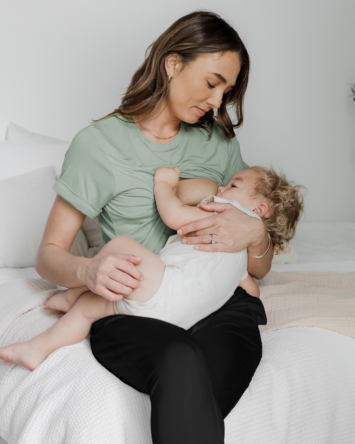 Model sitting on bed nursing baby and wearing the Everyday Asymmetrical Nursing T-shirt in Aqua Mist @model_info:Kate is wearing a Small.