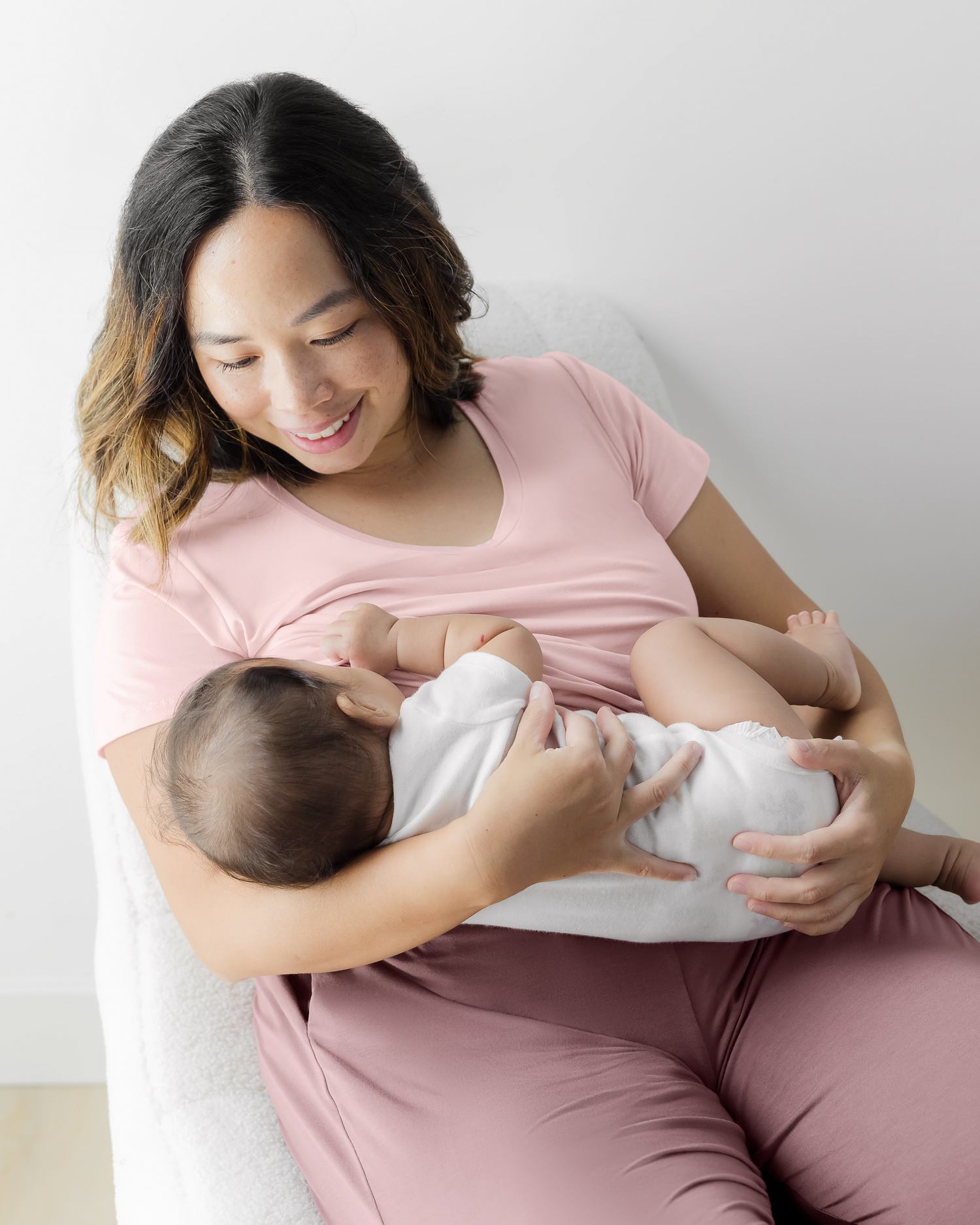 Model sitting in chair wearing the Everyday Maternity & Nursing T-shirt in Dusty Pink and nursing baby @model_info:Elizabeth is wearing a Large.