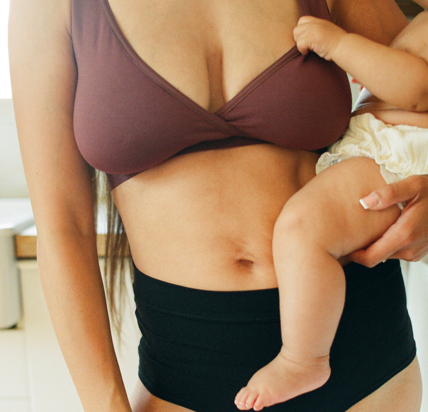 Model is holding baby and wearing Every Stage Crossover Bra in Cocoa and Every Stage Brief in Black with a kitchen background