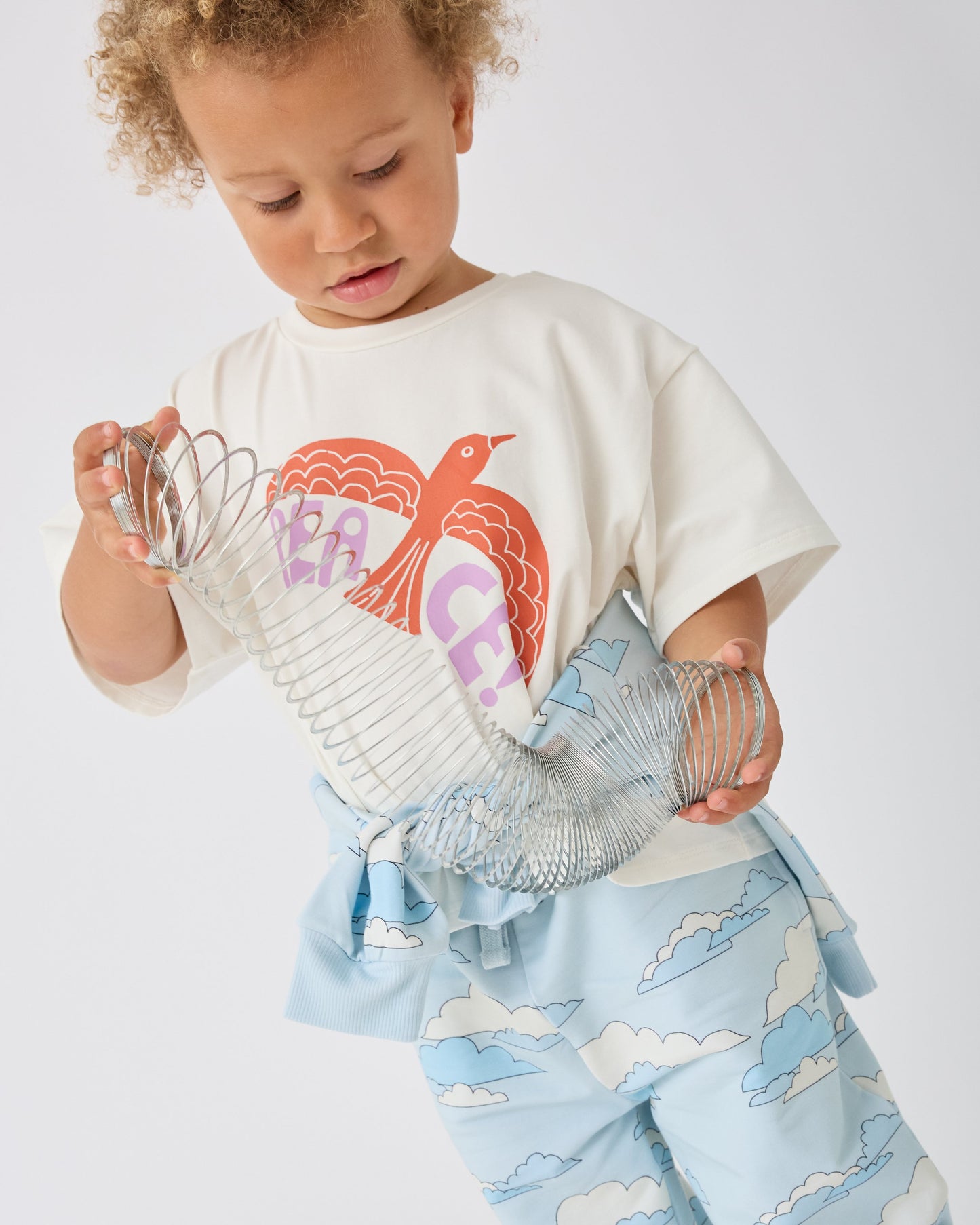 Model is wearing Graphic Tee in White Bird and French Terry Jogger in Blue Clouds while playing with a slinky, on a white background.@model_info:Giles is 2 years old and wearing 4T.