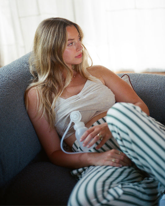 Model is wearing Sublime® Bamboo Hands-Free Pumping Lounge & Sleep Bra in Oatmeal Heather and Model is wearing Clea Bamboo Long Sleeve Pajama Set in Green Candy Stripe on a blue sofa with light filled curtains in the background.