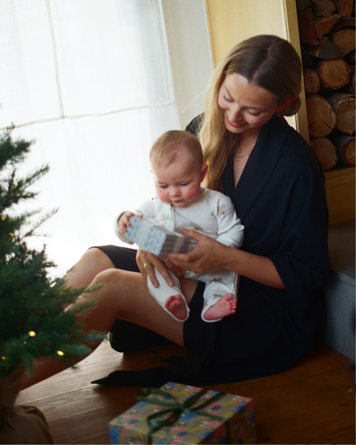 A woman in a dark navy robe sits on a wooden floor holding a baby dressed in Pointelle Baby Pajama in Ivory Bears, who is looking at a gift box. A wrapped gift box sits in the foreground, with a Christmas tree on the left and a stack of firewood in the background.