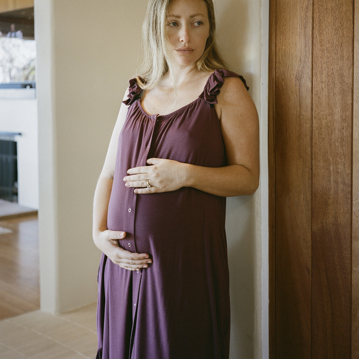 Model standing at in home while wearing Ruffle Strap Labor & Delivery Gown in Burgundy Plum.