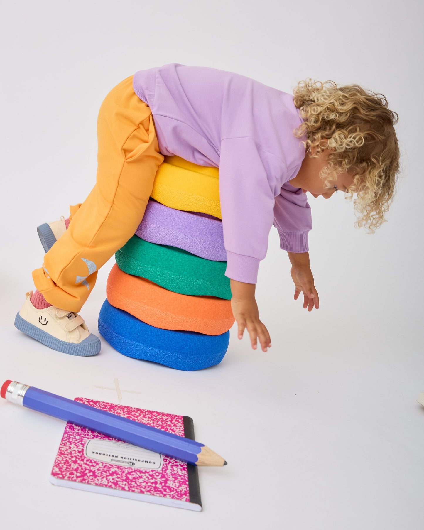 Model is wearing Graphic Sweatshirt in Purple Bird and French Terry Jogger in Orange Bird while playing on a stack of foam steps next to a giant pencil and notebook, on a gray background.
