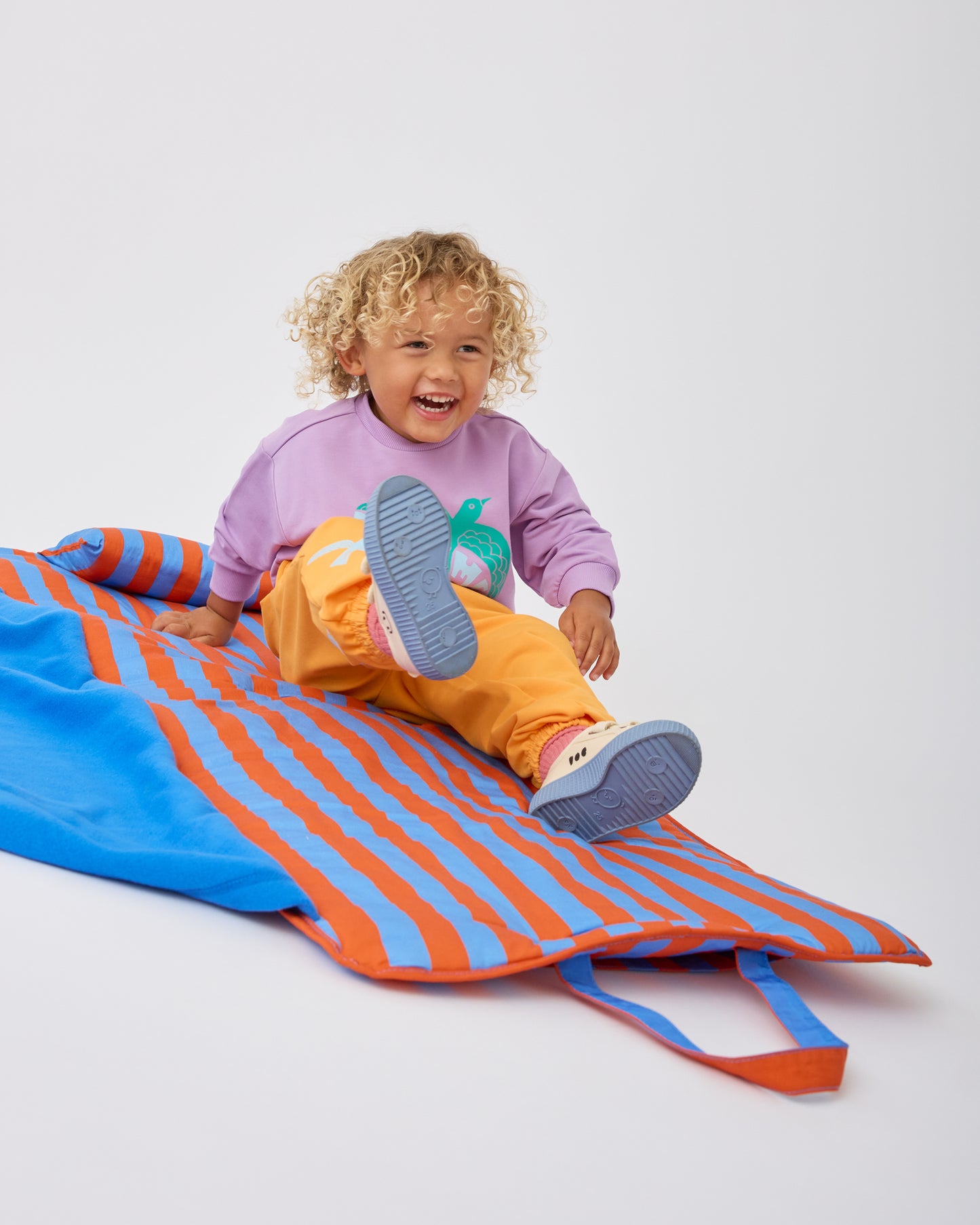 Child is sitting up and laughing on the Cool Kid Nap Mat in Blue & Rust Stripe on a beige background. 