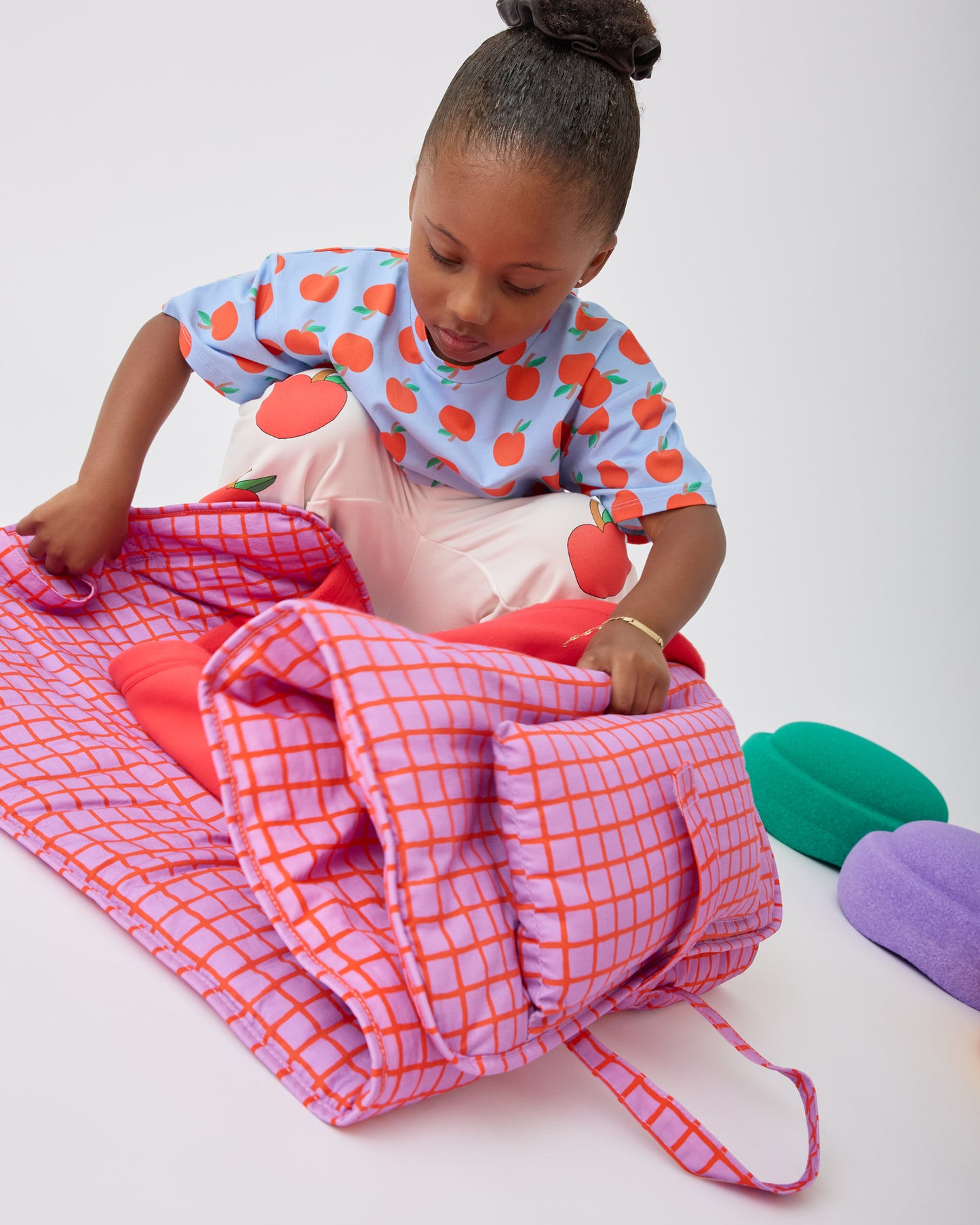 Child is unfolding her Cool Kid Nap Mat in Orchid Grid on a beige background.