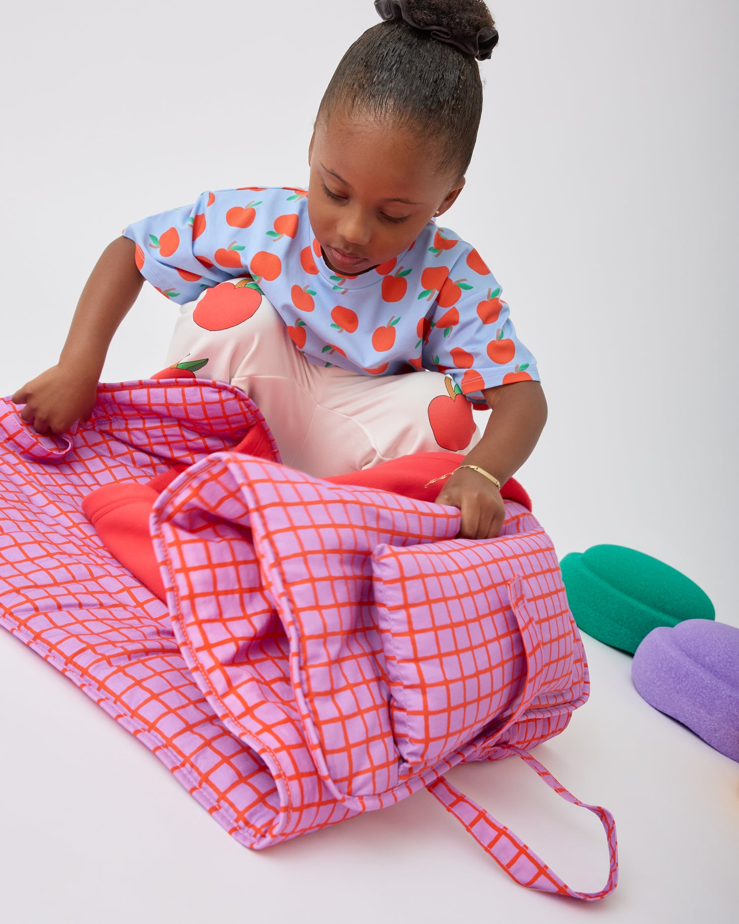 Child is unfolding her Cool Kid Nap Mat in Orchid Grid on a beige background.