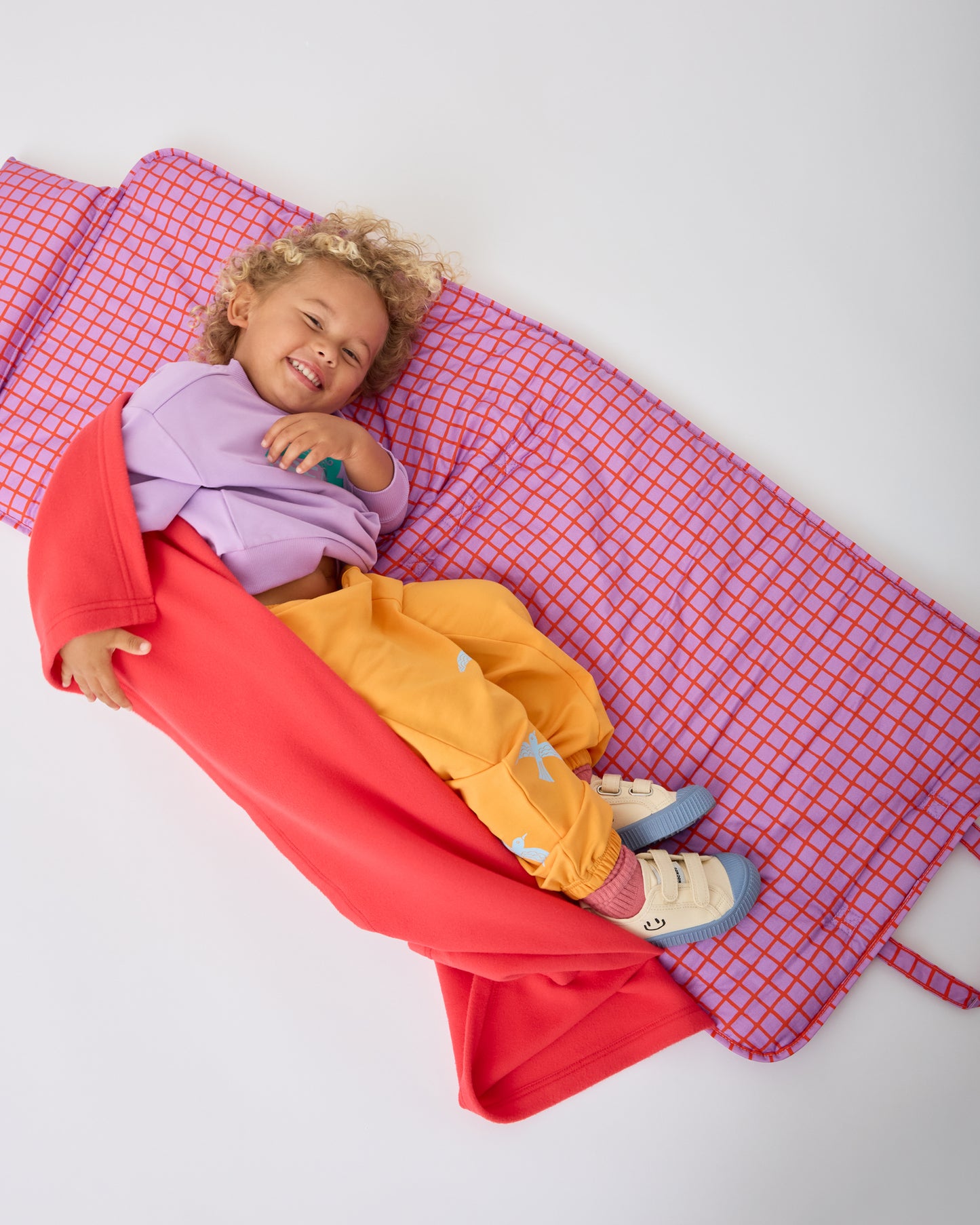 Child is smiling and rolling around on the Cool Kid Nap Mat in Orchid Grid on a beige background. @model_info:Waylon is 3 years old.
