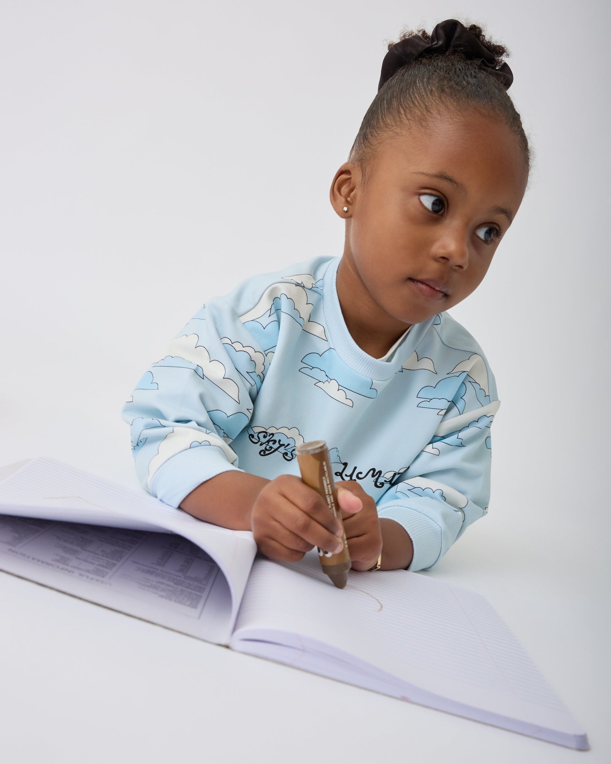 Model is wearing Graphic Sweatshirt in Blue Clouds while drawing in a sketch book, on a gray background.@model_info:Aliyana is 4 years old and wearing 4T.