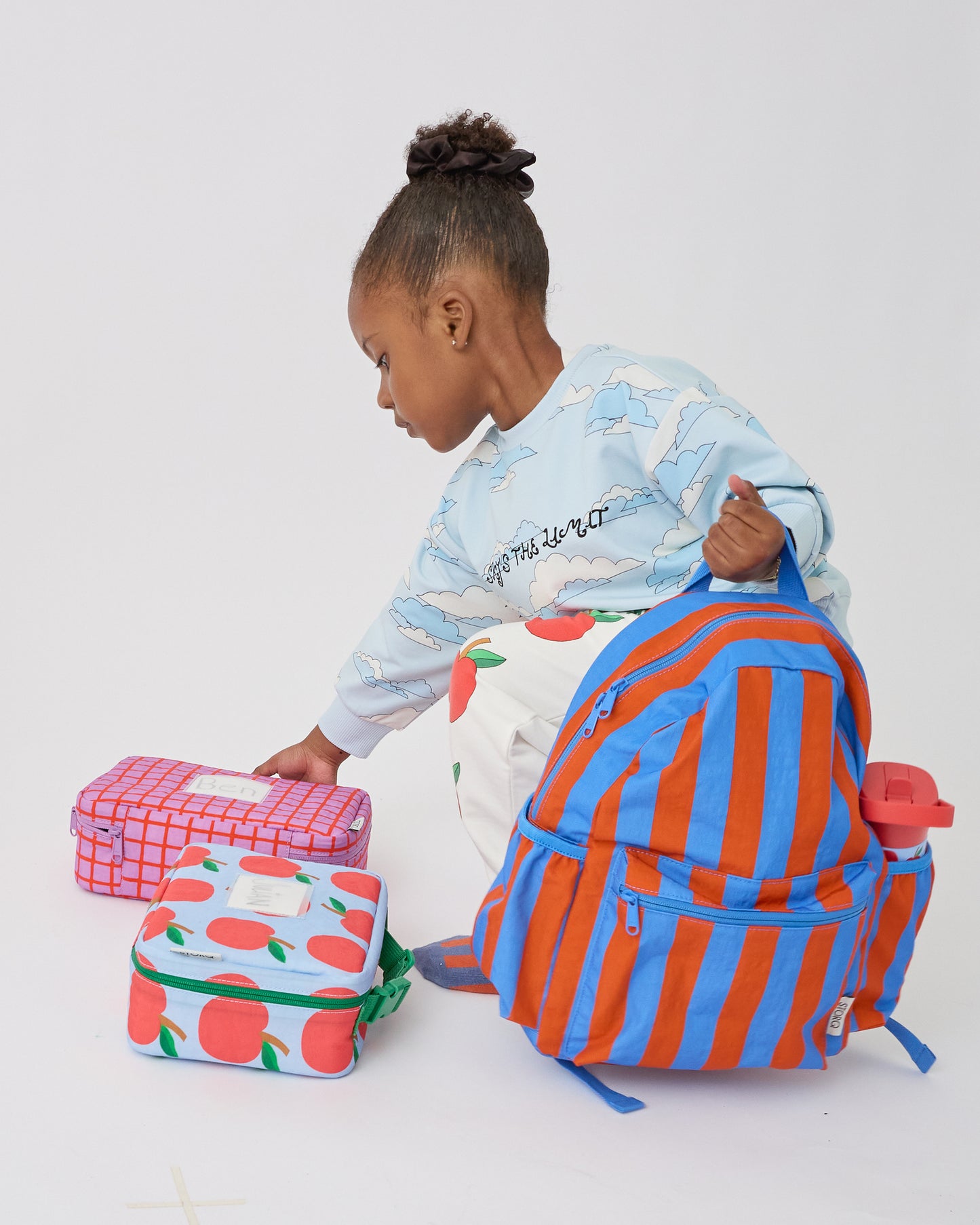 Child is holding the Cool Kid Backpack in Blue & Rust Stripe with Cool Kid Water Bottle in Blue Apple, Lunch Box in Blue Apple and Orchid Grid, on a beige background. @model_info:Aliyana is 4 years old.