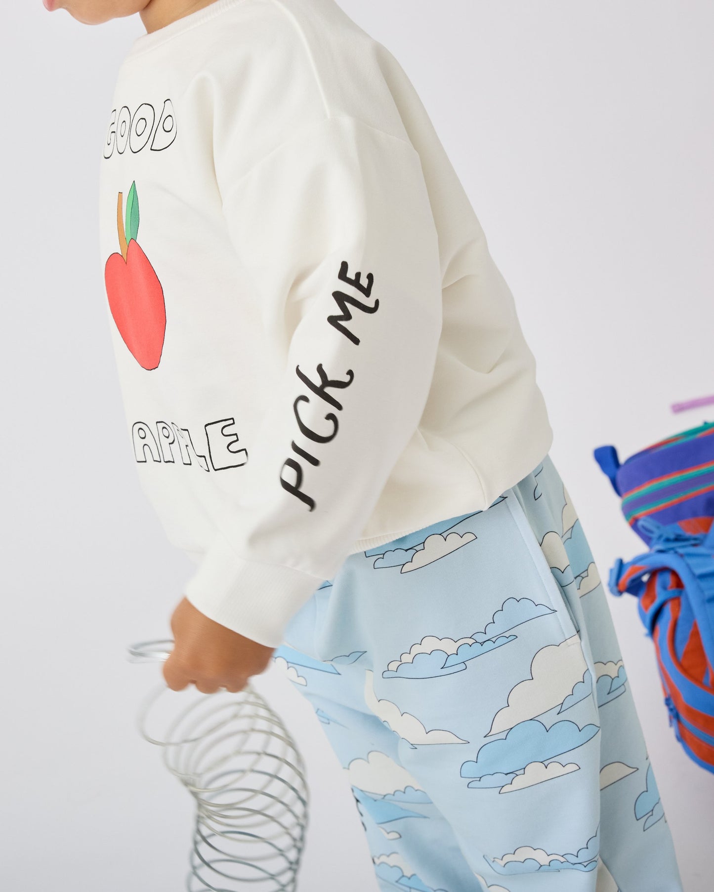 Model is wearing Graphic Sweatshirt in Ivory Apple and French Terry Jogger in Blue Clouds while playing with a slinky and a backpack in the background on a white background.