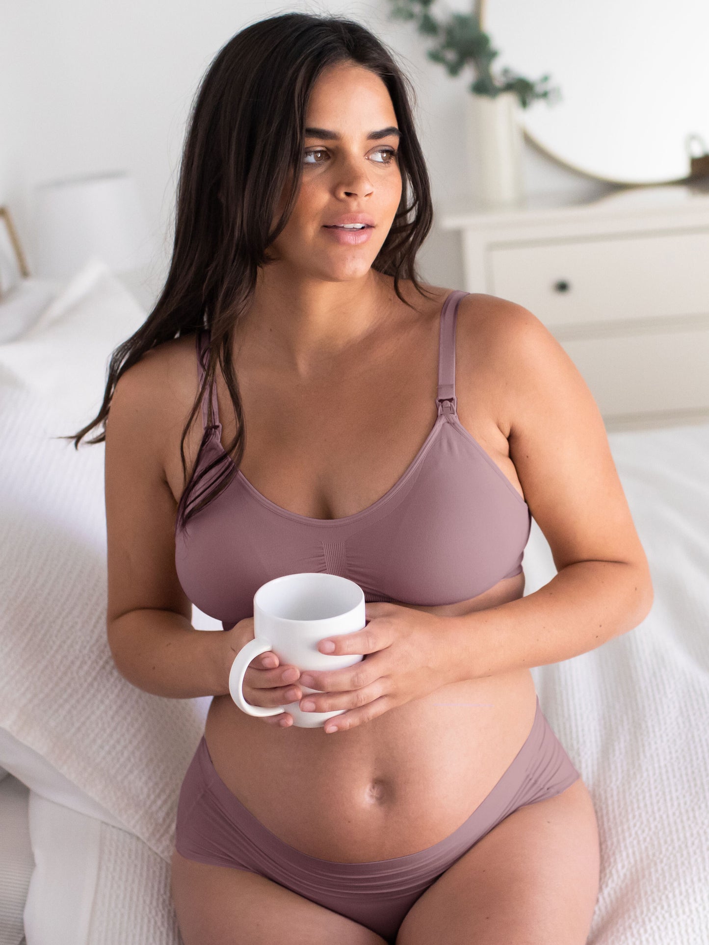 A pregnant model wearing the Simply Sublime® Nursing Bra in twilight sitting on the edge of the bed