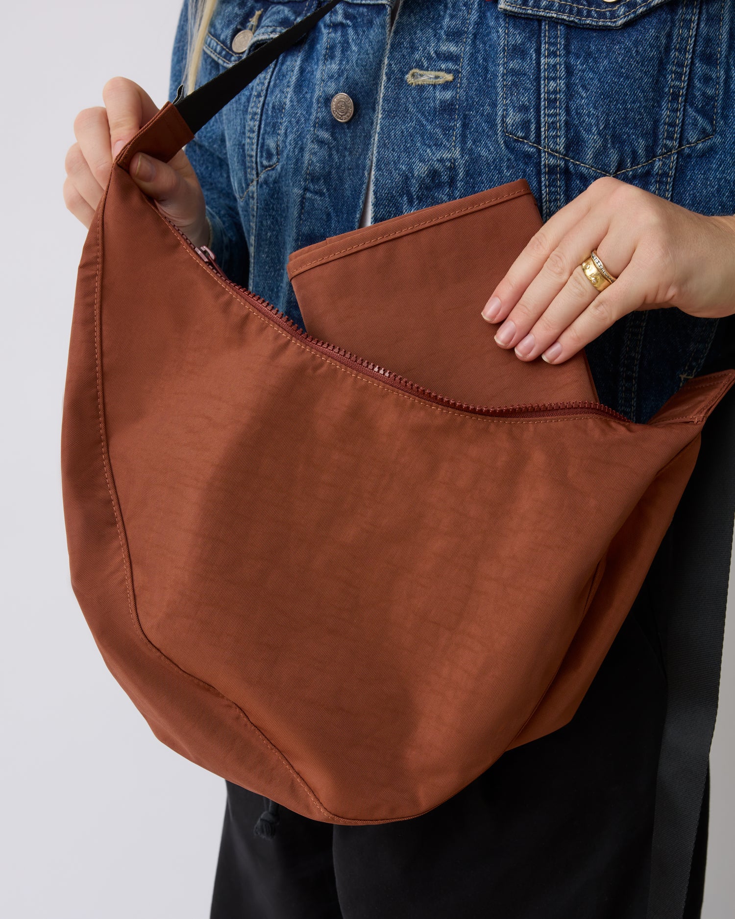 Model is wearing Half Moon Crossbody Bag in Cinnamon while placing paired diaper changing mat back inside, on a beige background. 