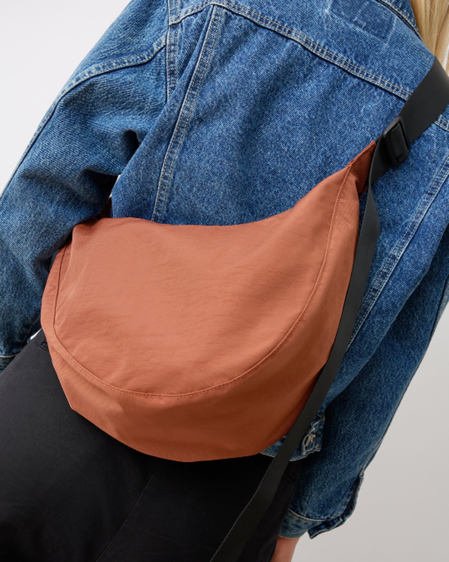 Model is wearing Half Moon Crossbody Bag in Cinnamon, across her back, on a beige background.