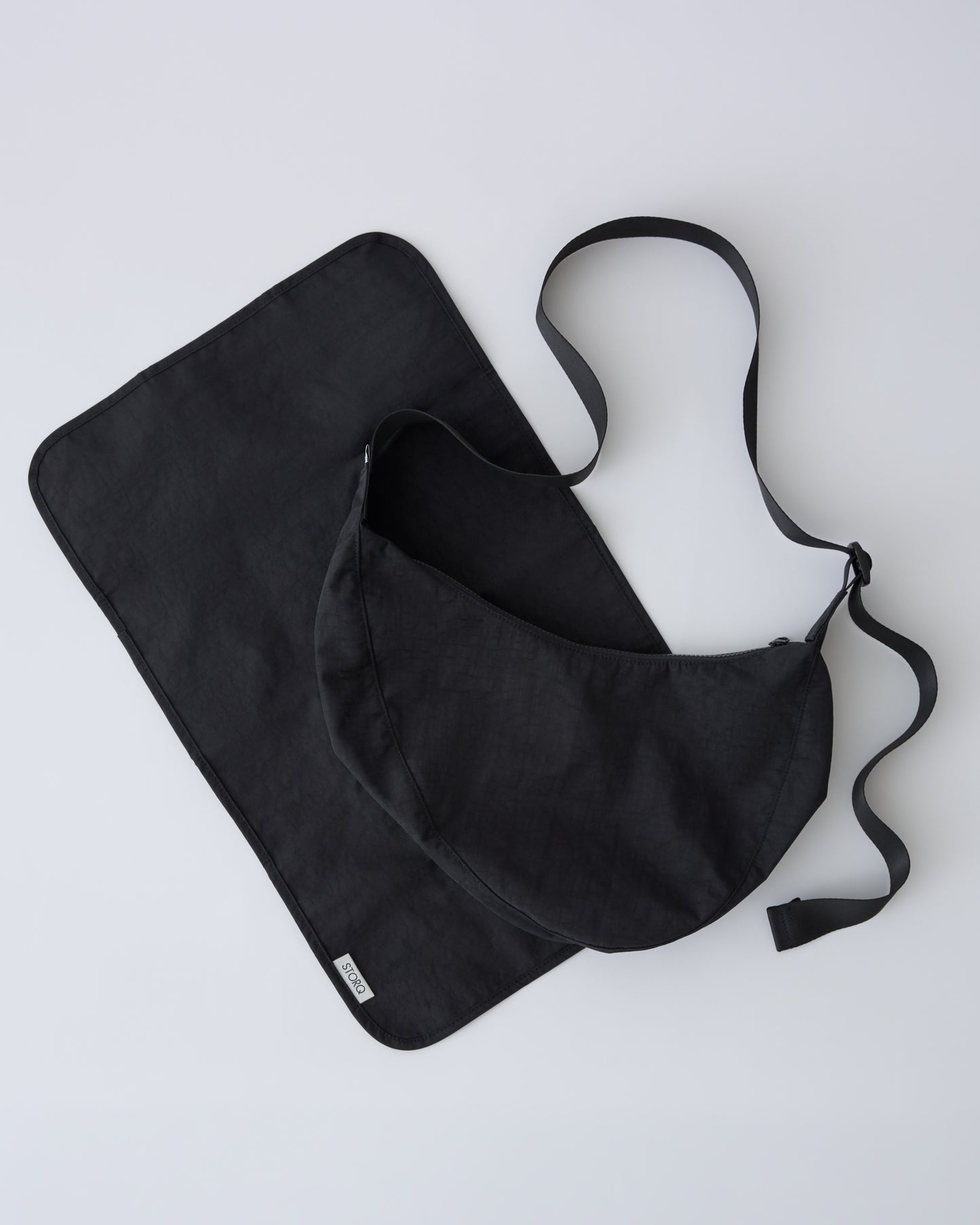 Half Moon Crossbody Bag in Black with paired diaper changing mat on a beige background. 