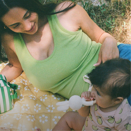 Mom laying on Inside/Outside Play Mat in Sunshine Dots and wearing Ribbed Bamboo Scoop Neck Nursing Tank in Avocado next to baby wearing Bamboo Short Sleeve & Shorts Set in Pink Pomelo on a grassy patch. Next to mom is Mini Cooler Bag in Green Stripe.@model_info:Zoe is wearing a Small.