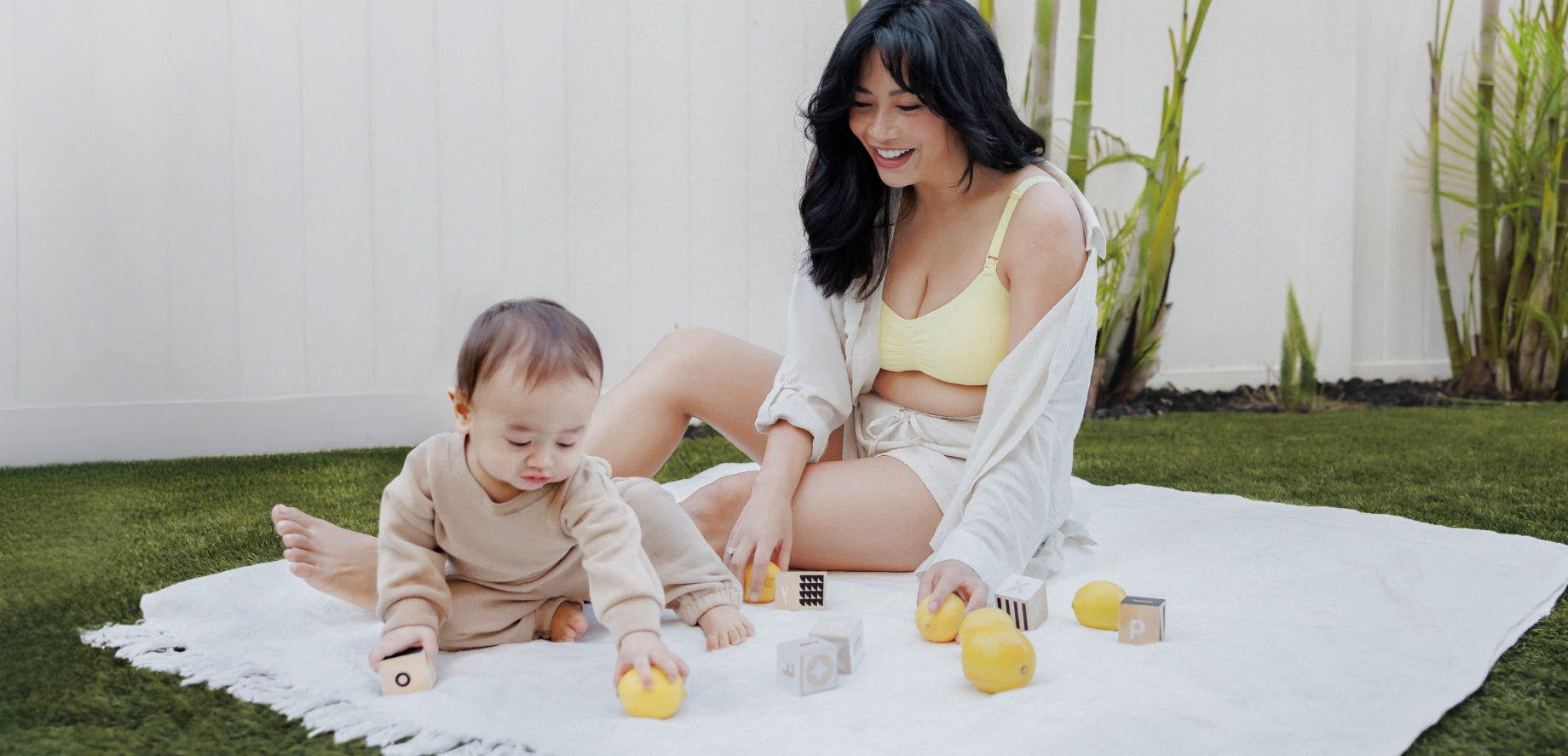 Model sitting on blanket on grass with baby wearing the Sublime® Hands-Free Pumping & Nursing Bra in Light Lemon, paired with the Reese Oversized Linen Button Down Shirt and the Reese Linen Short in Natural