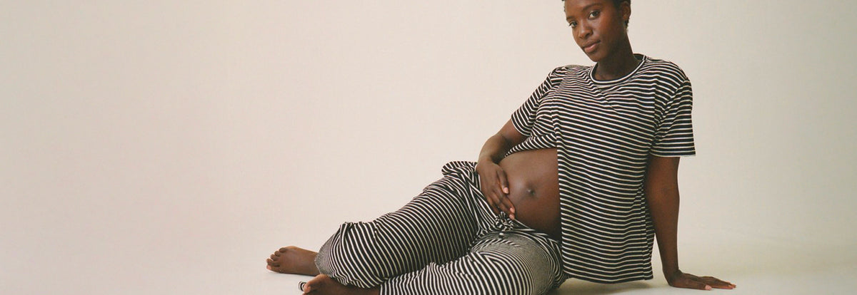 Model is showing baby bump and wearing Luxe Terry Maternity & Nursing Split Hem T-shirt in Black & Ivory Stripe and Luxe Terry Maternity & Postpartum Pant in Black & Ivory Stripe, on a beige background.
