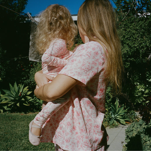 Mother and daughter wearing matching pajamas in their sunny, lush yard. Woman is wearing Cotton Bamboo Pajama Set in Pink Toile and daughter is wearing Bamboo Baby Pajama in Pink Toile and soft ballet shoes.