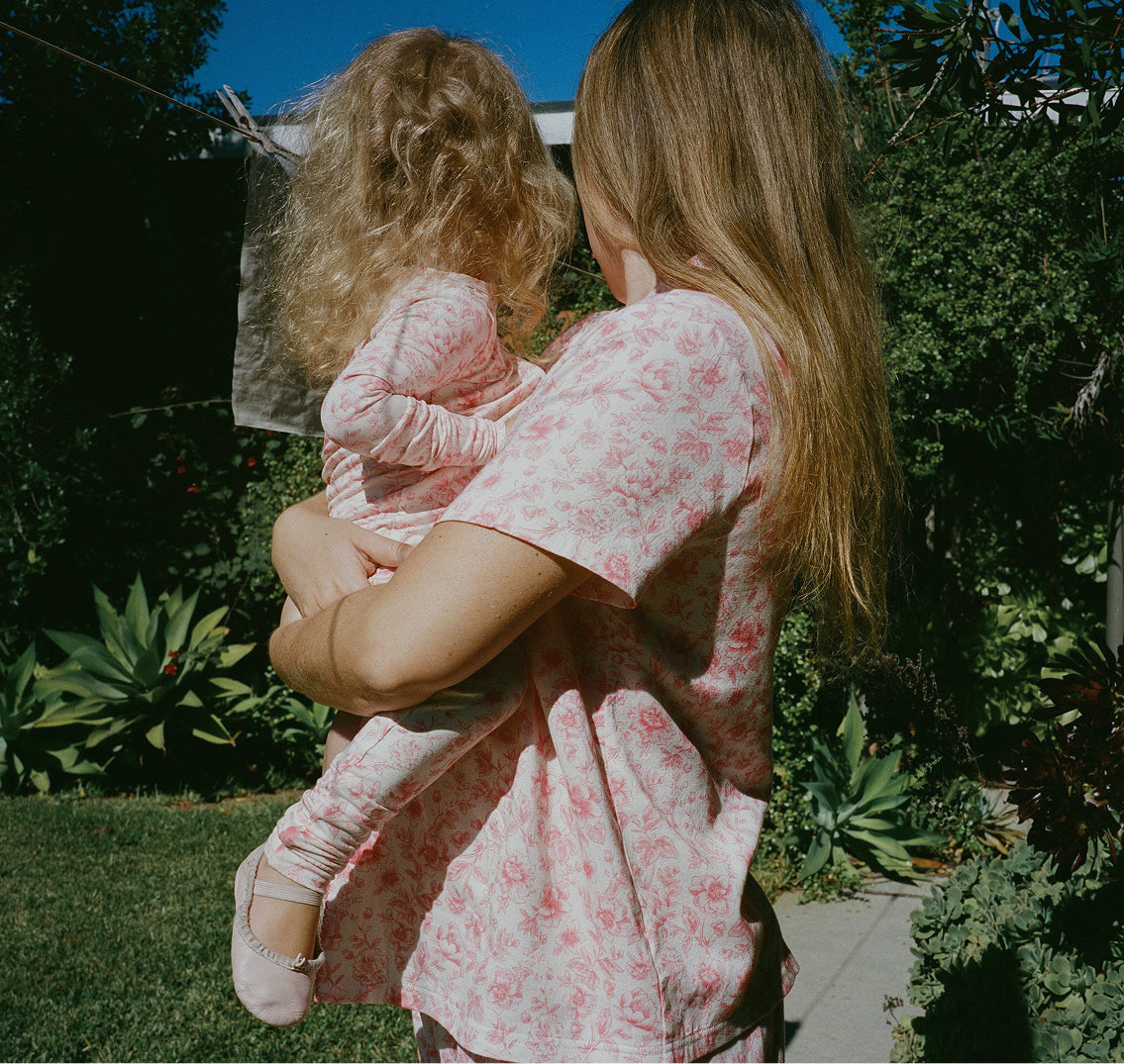 Mother and daughter wearing matching pajamas in their sunny, lush yard. Woman is wearing Cotton Bamboo Pajama Set in Pink Toile and daughter is wearing Bamboo Baby Pajama in Pink Toile and soft ballet shoes.
