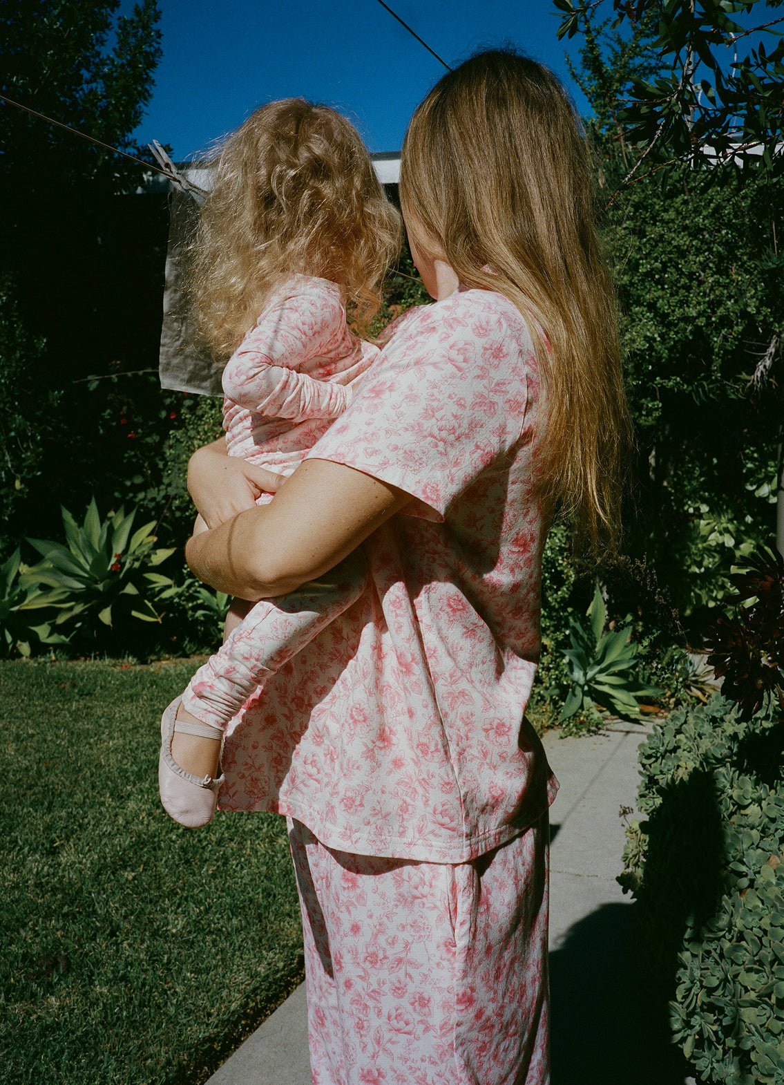 Mother and daughter wearing matching pajamas in their sunny, lush yard. Woman is wearing Cotton Bamboo Pajama Set in Pink Toile and daughter is wearing Bamboo Baby Pajama in Pink Toile and soft ballet shoes.