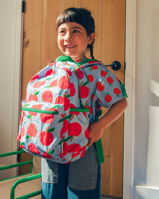 Smiling girl standing in a light filled room while wearing the Cool Kid Backpack in Blue Apple backwards.