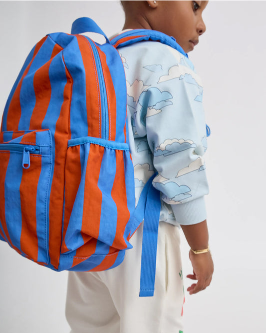 Child is wearing Cool Kid Backpack in Blue & Rust Stripe, on a beige background.