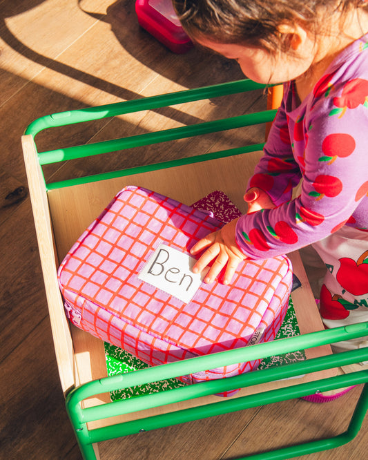 Child is reaching for Cool Kid Lunch Box in Orchid Grid with name tag in clear pocket, next to journals on a chair in a sun filled room.