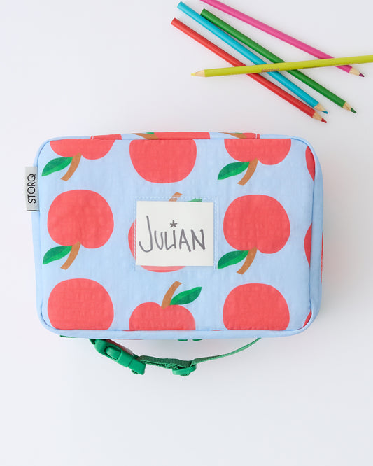 Cool Kid Lunch Box in Blue Apple with name tag in card pocket, next to coloring pencils on a beige background.