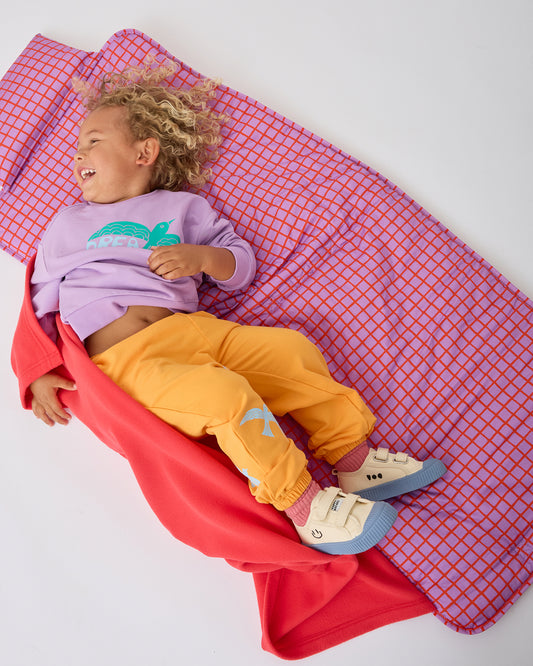 Child is smiling and rolling around on the Cool Kid Nap Mat in Orchid Grid on a beige background. 
